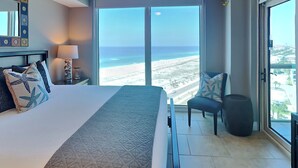 A hotel room with a large bed, a chair, a lamp, and a view of the beach. - Portofino Island Resort 5 by Southern Vacation Rentals (Pensacola Beach)