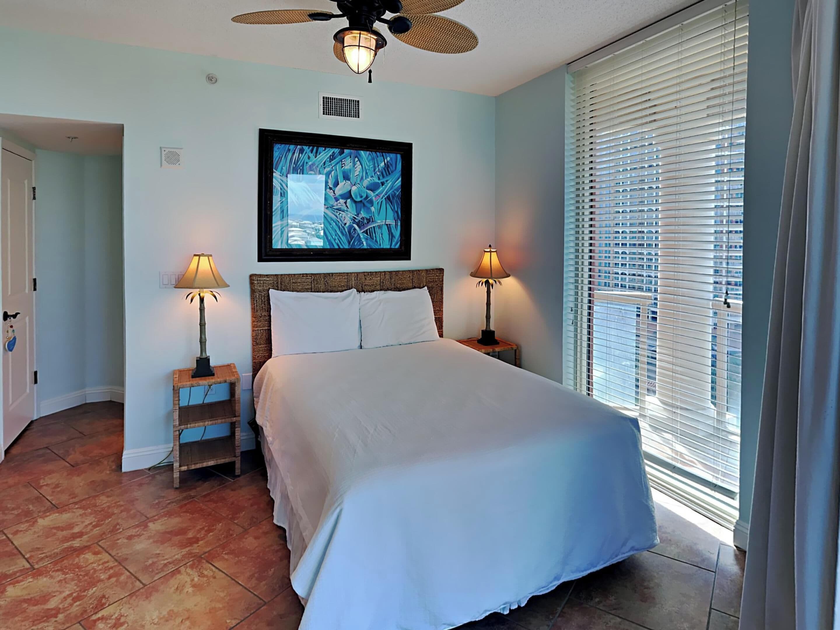 A bedroom with a bed, two bedside lamps, a ceiling fan, a framed artwork, and a window with blinds.