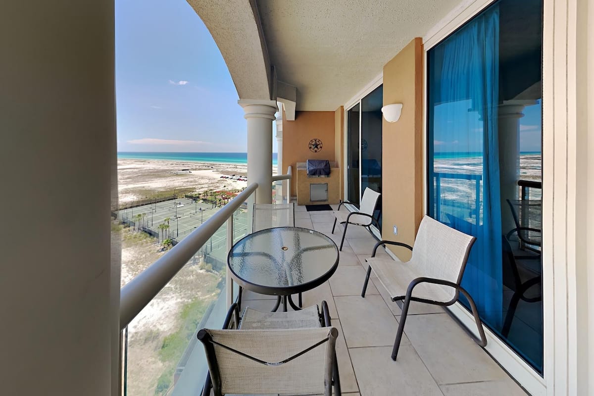 Een balkon met een glazen tafel en stoelen, met uitzicht op een strand en de oceaan.