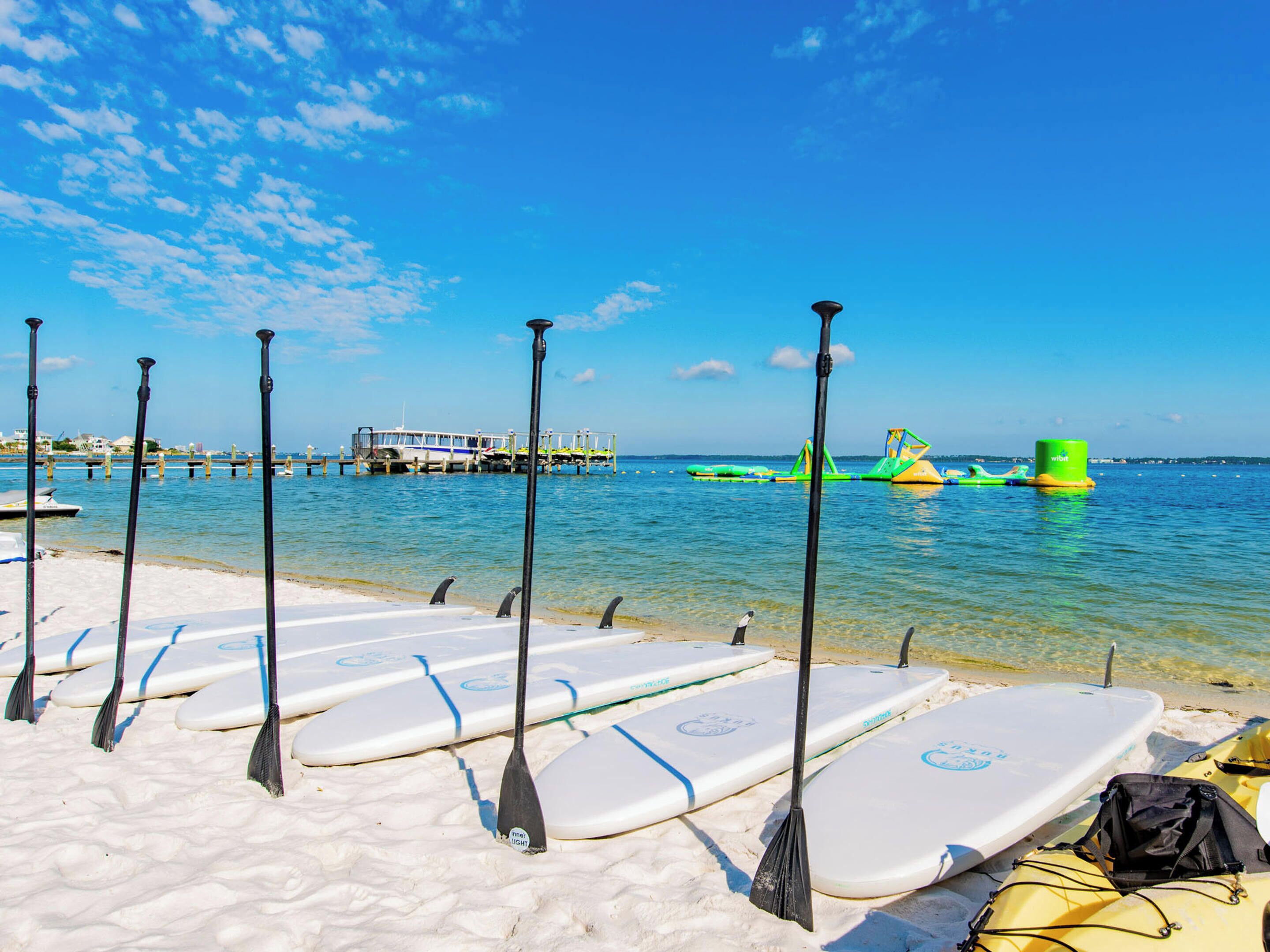 A beach with paddleboards and paddles lined up on the sand, a pier in the background, and clear blue skies.