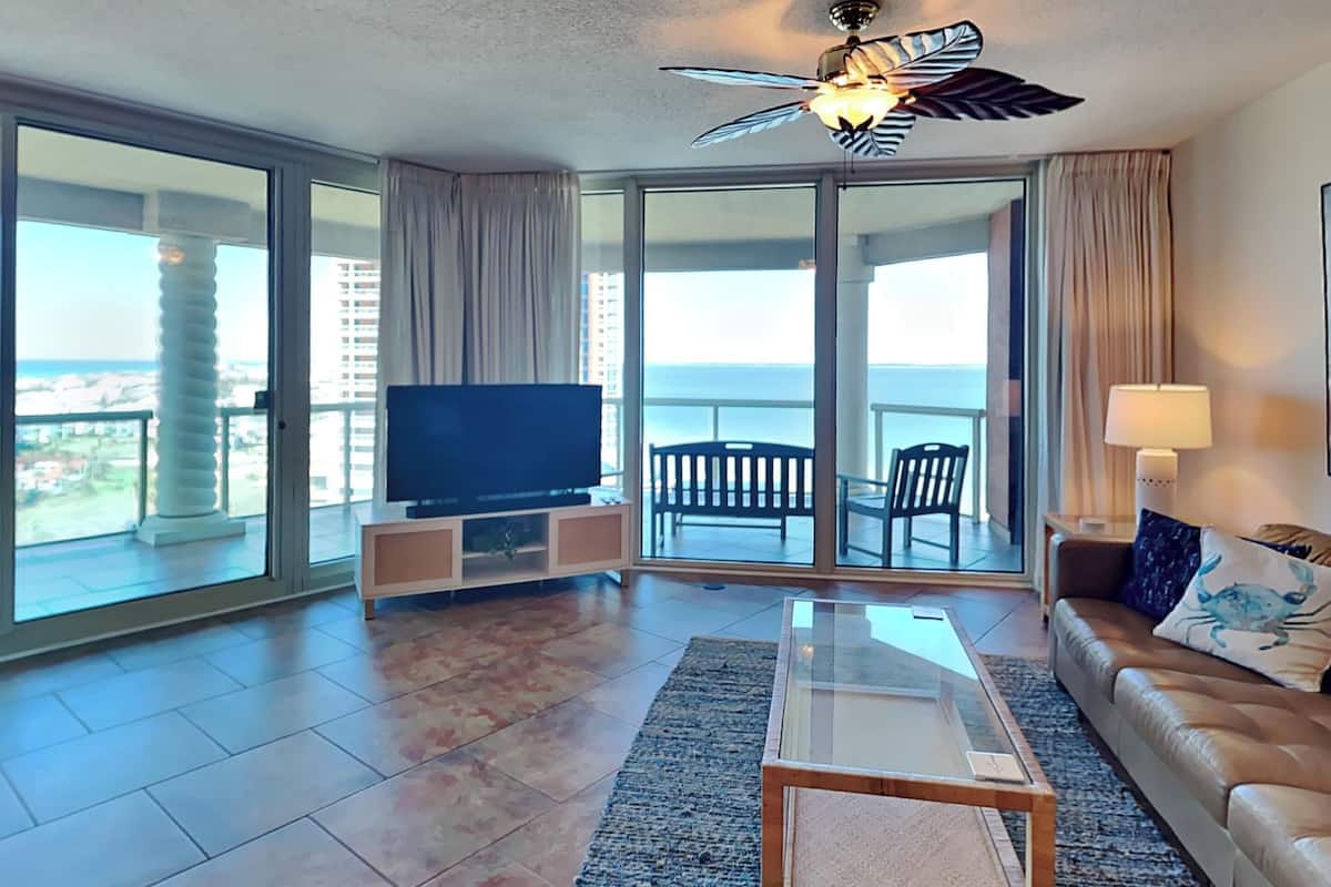 Een ruime woonkamer met een grote flatscreen televisie, een glazen salontafel en een leren bank. De kamer heeft een balkon met uitzicht op de stad en een plafondventilator.