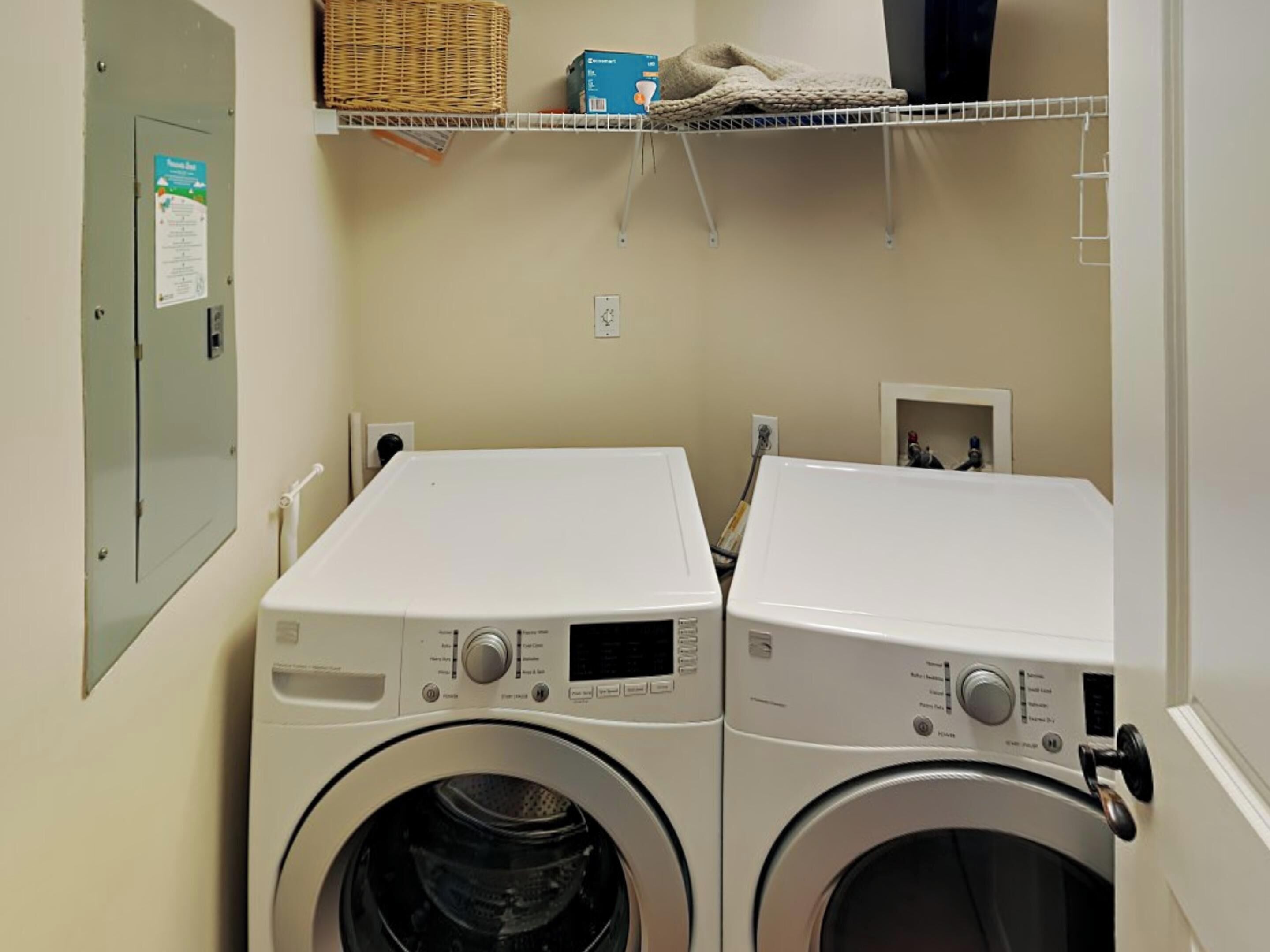 A laundry area with two front-loading washing machines, a wicker basket, a box, and a small shelf with a basket and a blanket.