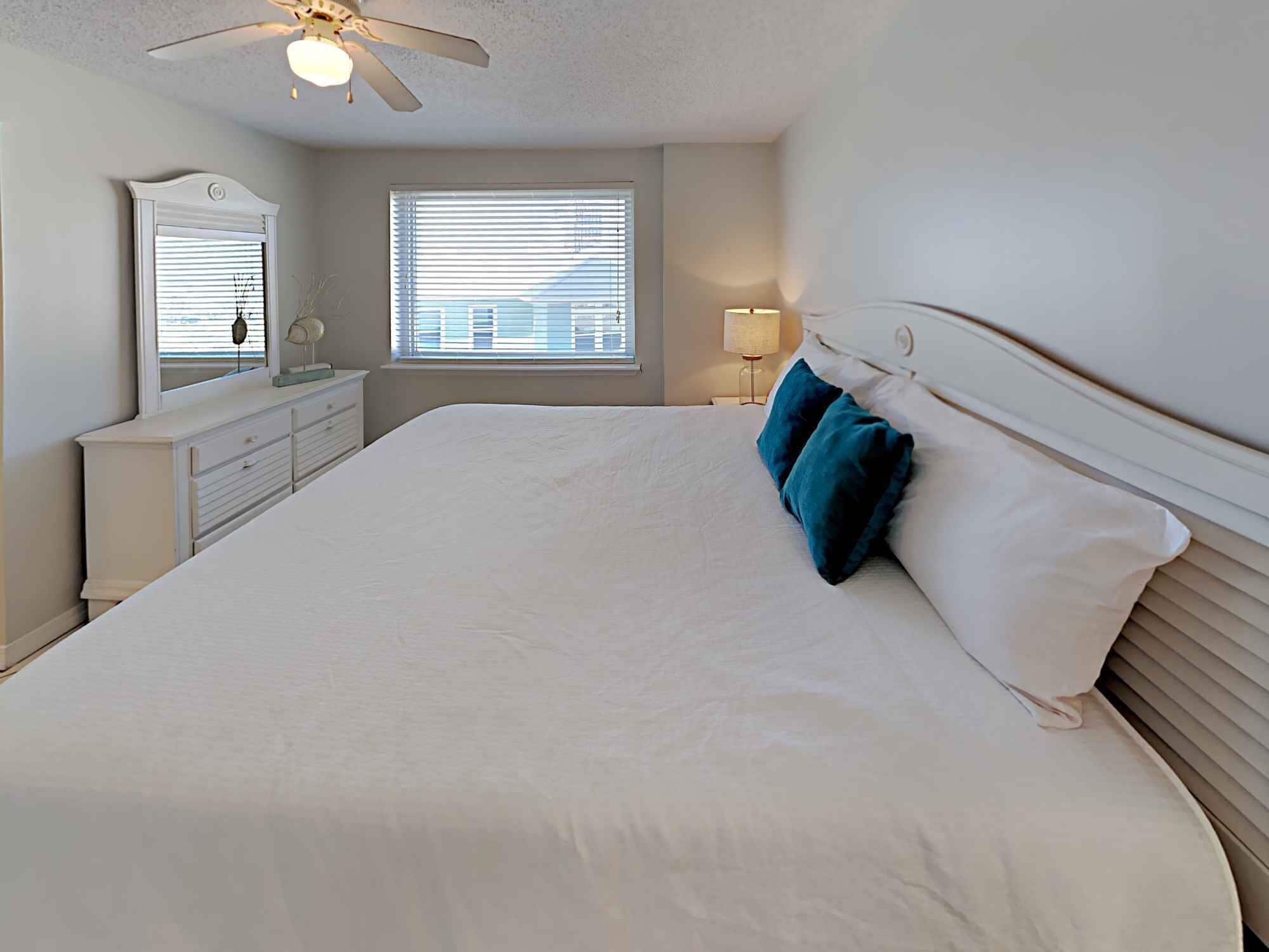 A bedroom with a large bed, a dresser, a mirror, a ceiling fan, and a window with blinds.
