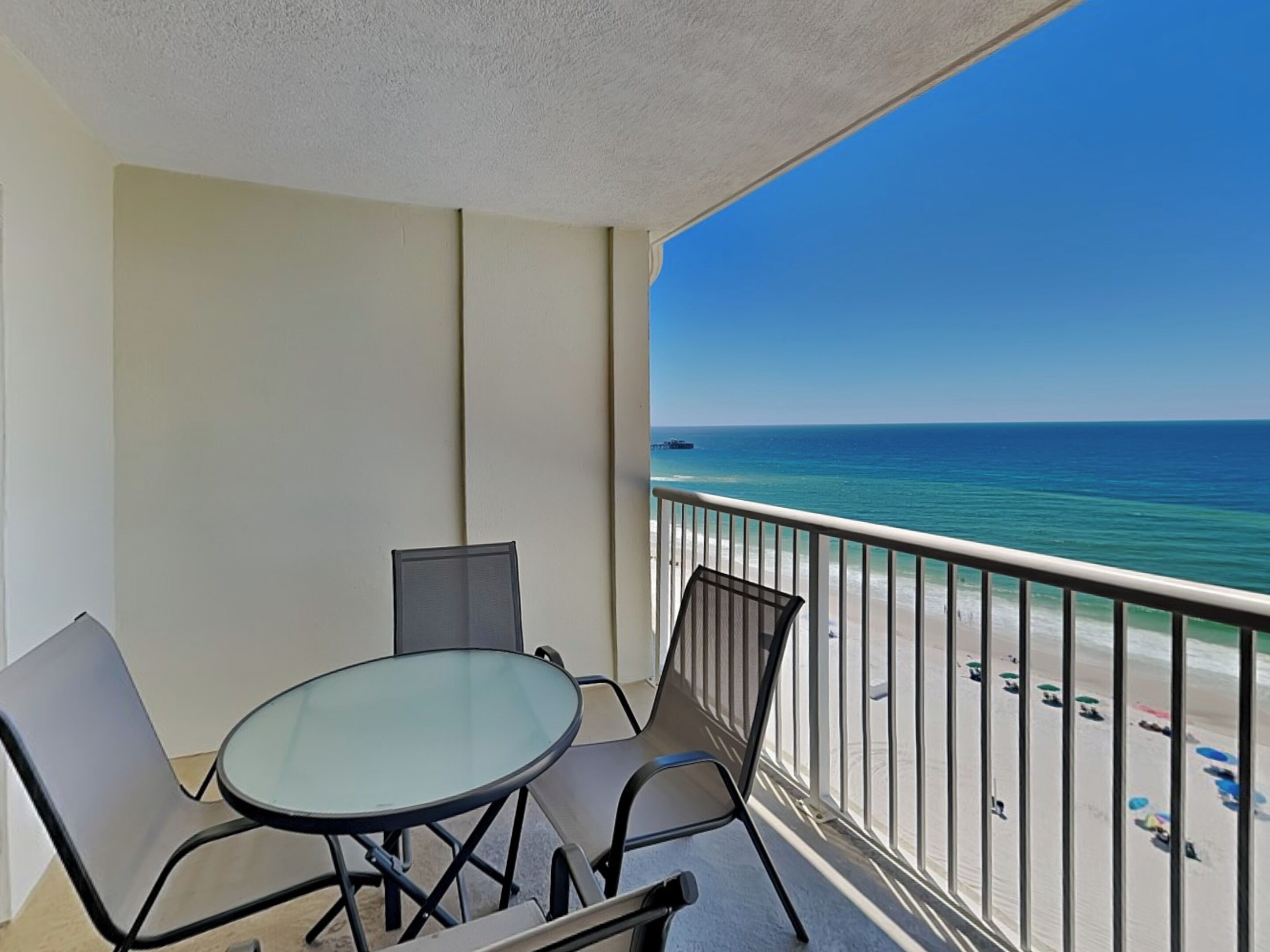 A balcony with a glass table and chairs, overlooking a beach with clear blue water and a pier in the distance.