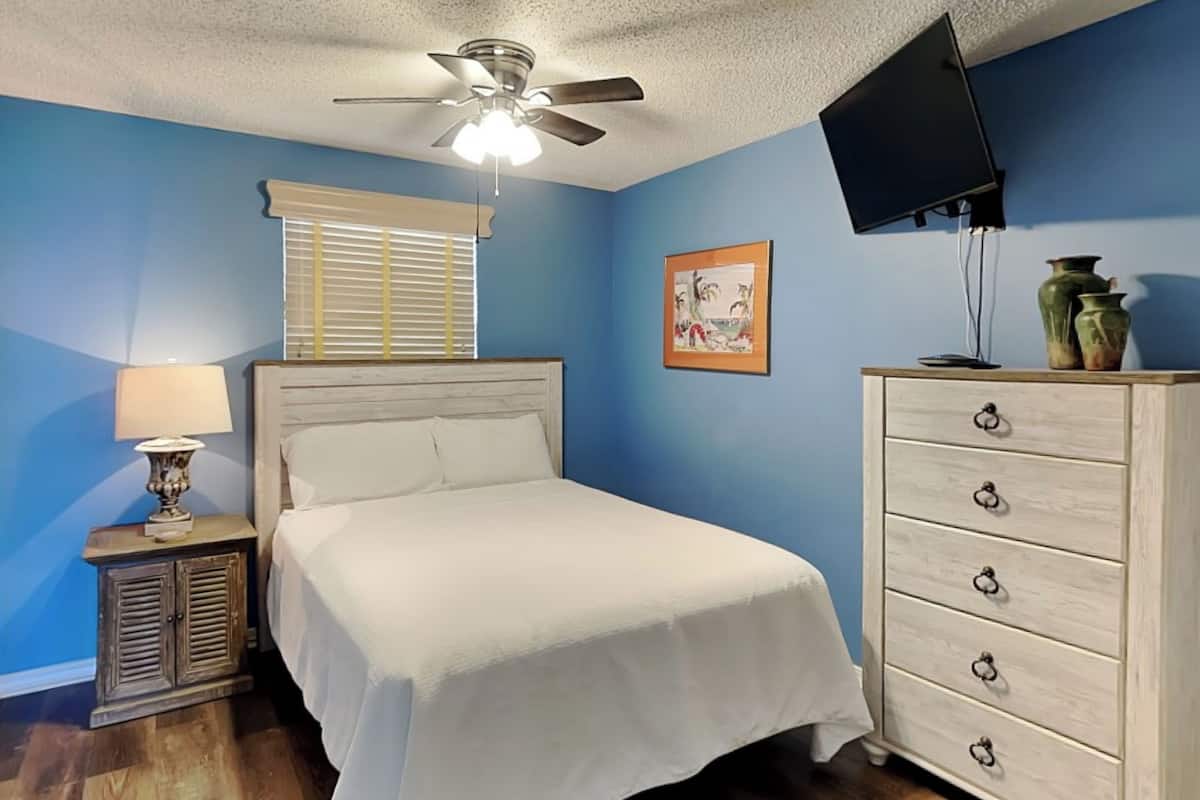 Un dormitorio con cama, mesita de noche con lámpara, cómoda, ventilador de techo y ventana con persianas, además de un cuadro en la pared.