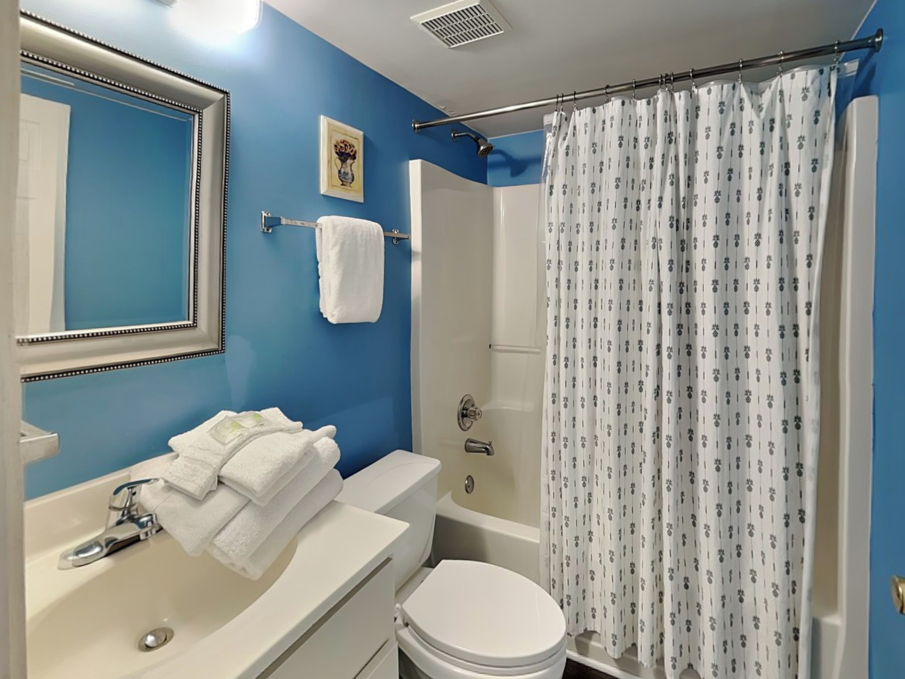 Ein Badezimmer mit blauer Wand, Waschbecken in Weiß, Spiegel, Handtuchhalter, Duschvorhang und Toilette.