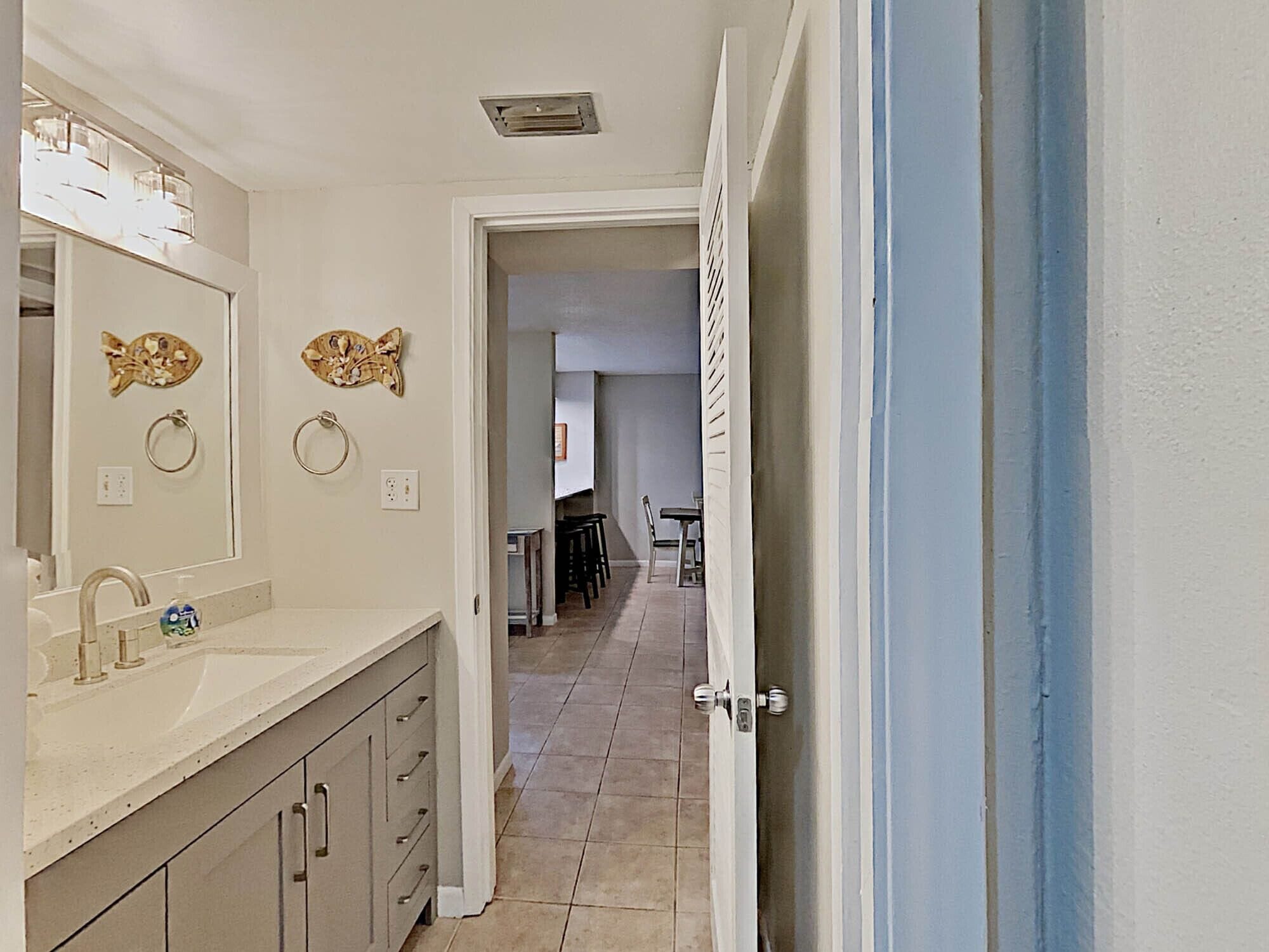 A narrow hallway with a bathroom on the left, featuring a sink and mirror, and a dining area with chairs and a table in the background.