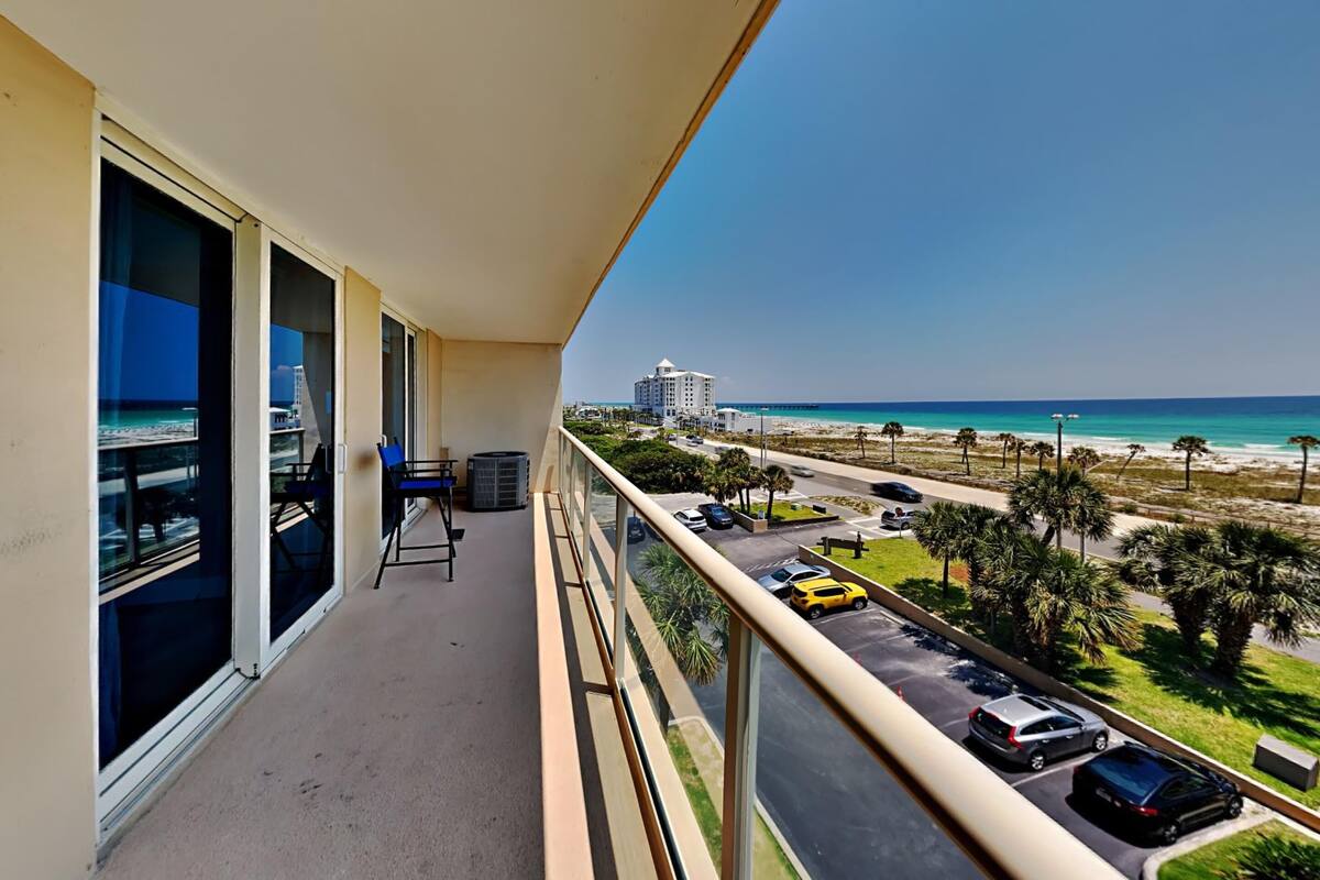 A balcony with a view of the beach, parked cars, and a distant building.
