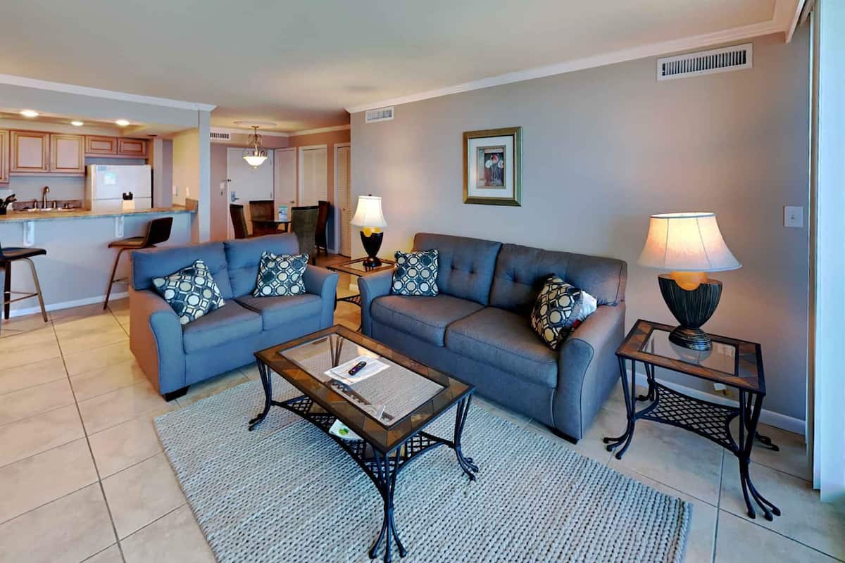 A living room with two sofas, a coffee table, and two side tables with lamps. There is a kitchen in the background.