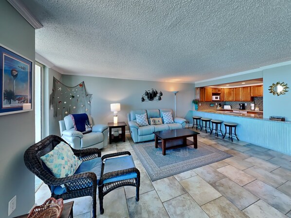 A living room with a blue sofa, wicker chairs, a coffee table, and a kitchen area with wooden cabinets and a bar counter. - Sabine Yacht & Racquet by Southern Vacation Rentals (Pensacola Beach)
