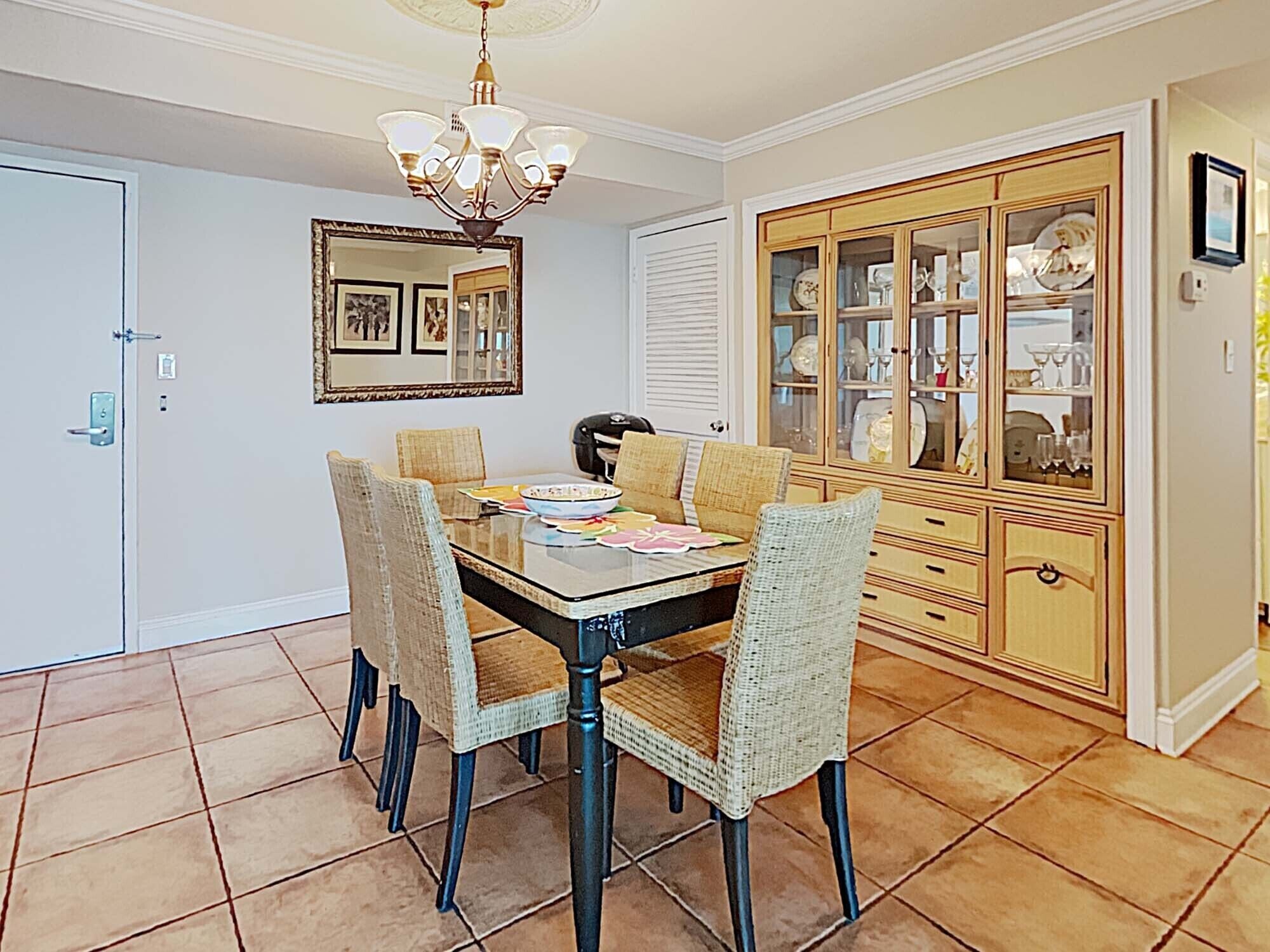 A dining room with a glass-top table, wicker chairs, a chandelier, a mirror, and a china cabinet.