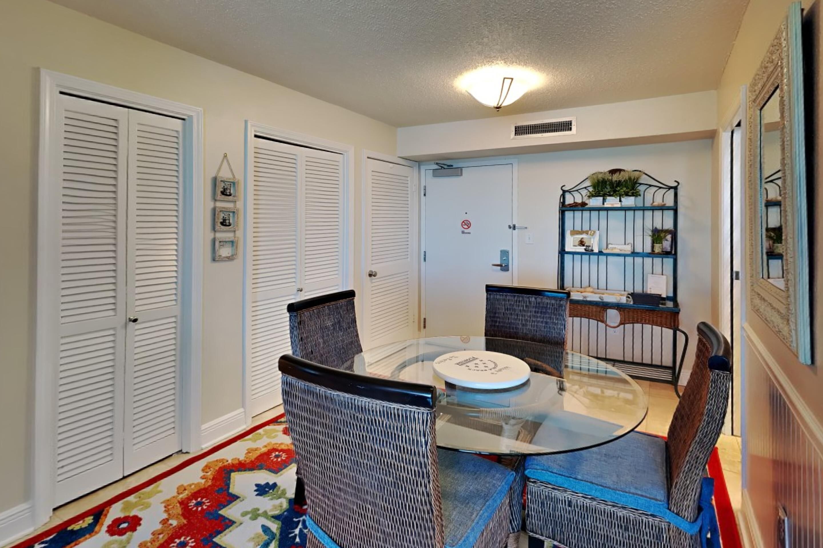 A dining area with a glass table and wicker chairs, white doors, and a small shelf with plants.