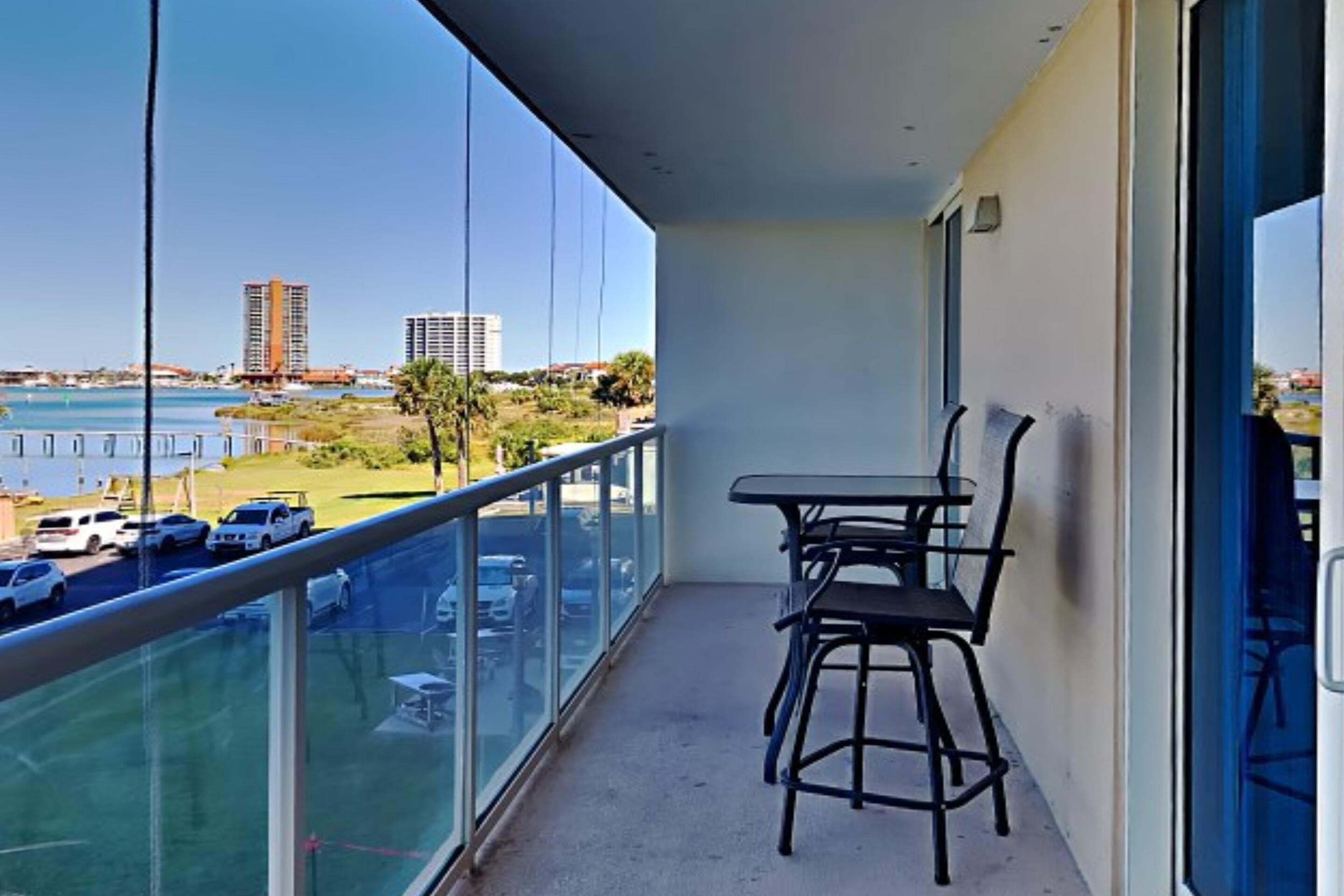 Un balcon avec une table et des chaises, donnant sur un parking et sur une étendue d’eau, avec des bâtiments visibles au loin.