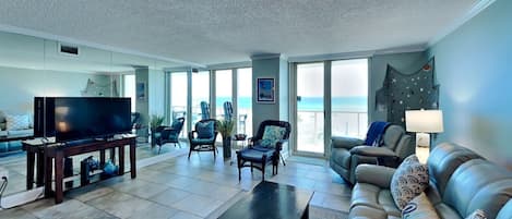 A spacious living room with a large flat-screen TV, comfortable seating, and a view of the ocean through large sliding doors.