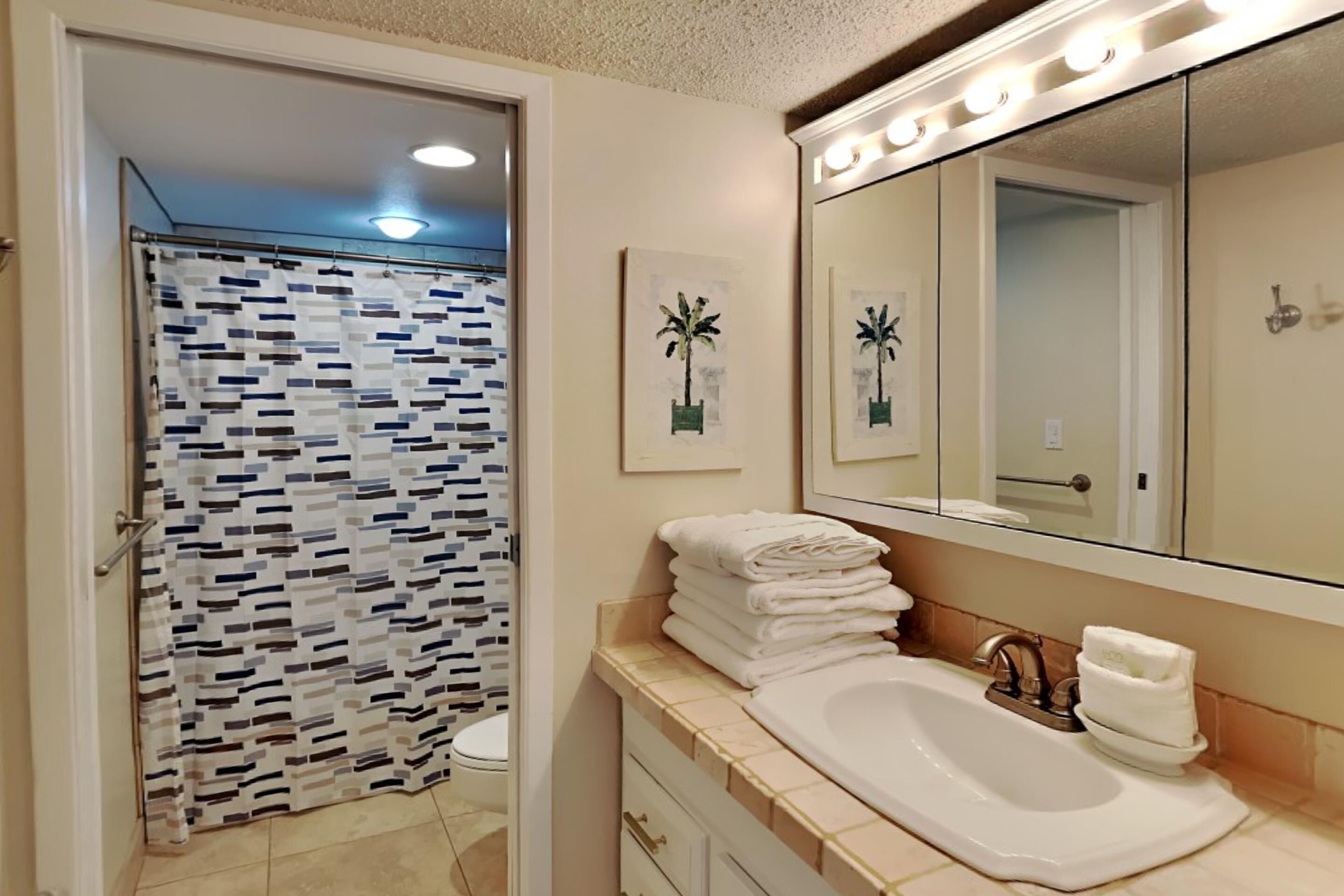 A bathroom with a shower curtain, a sink with a mirror, and a stack of towels.