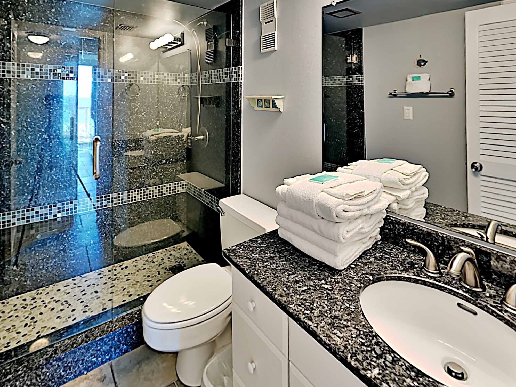 Un baño moderno con ducha de vidrio, inodoro blanco, lavamanos con encimera de granito y espejo.
