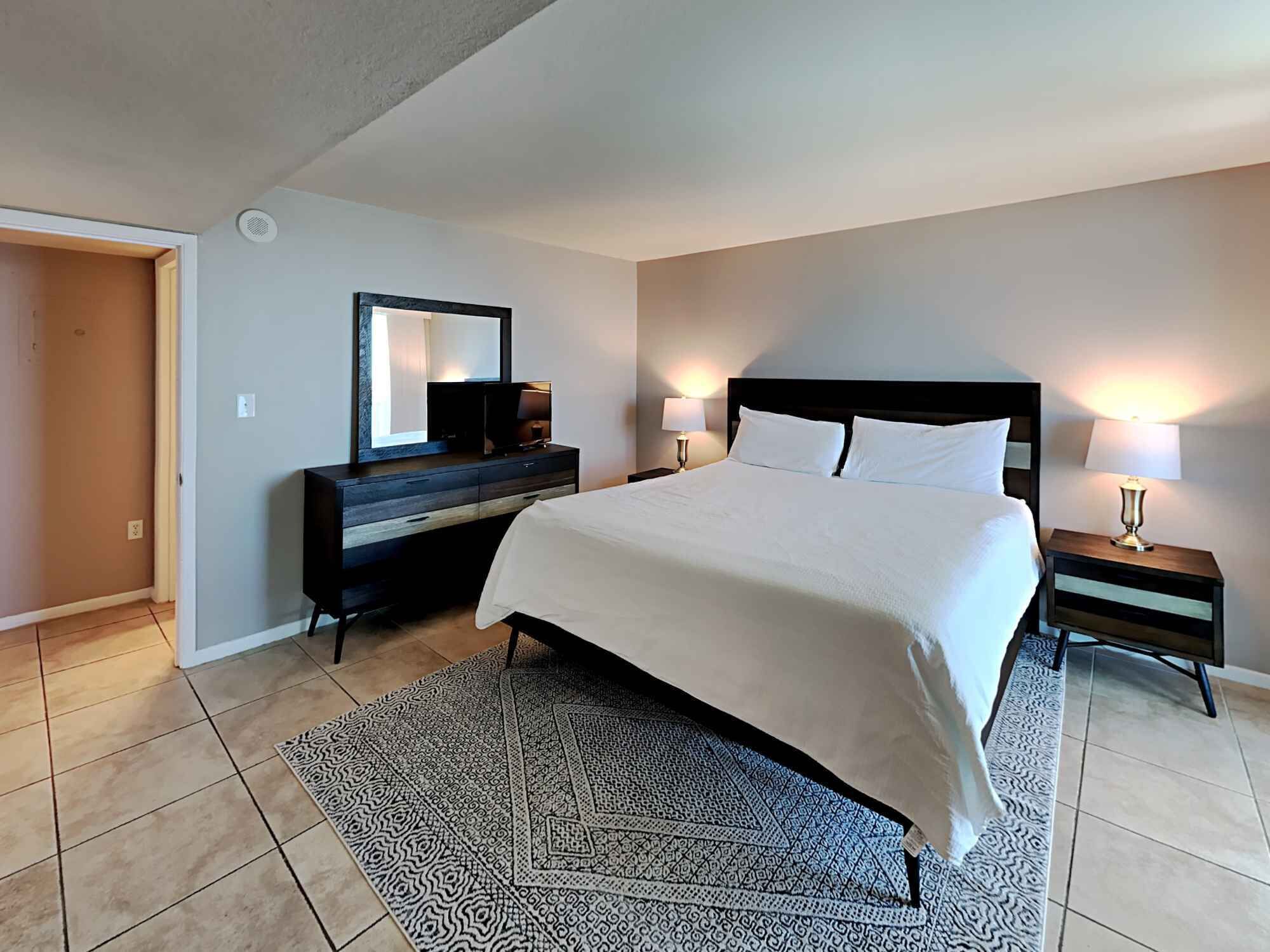 Une chambre d’hôtel avec un grand lit, une commode, un miroir, deux tables de chevet avec des lampes et un tapis à motifs.