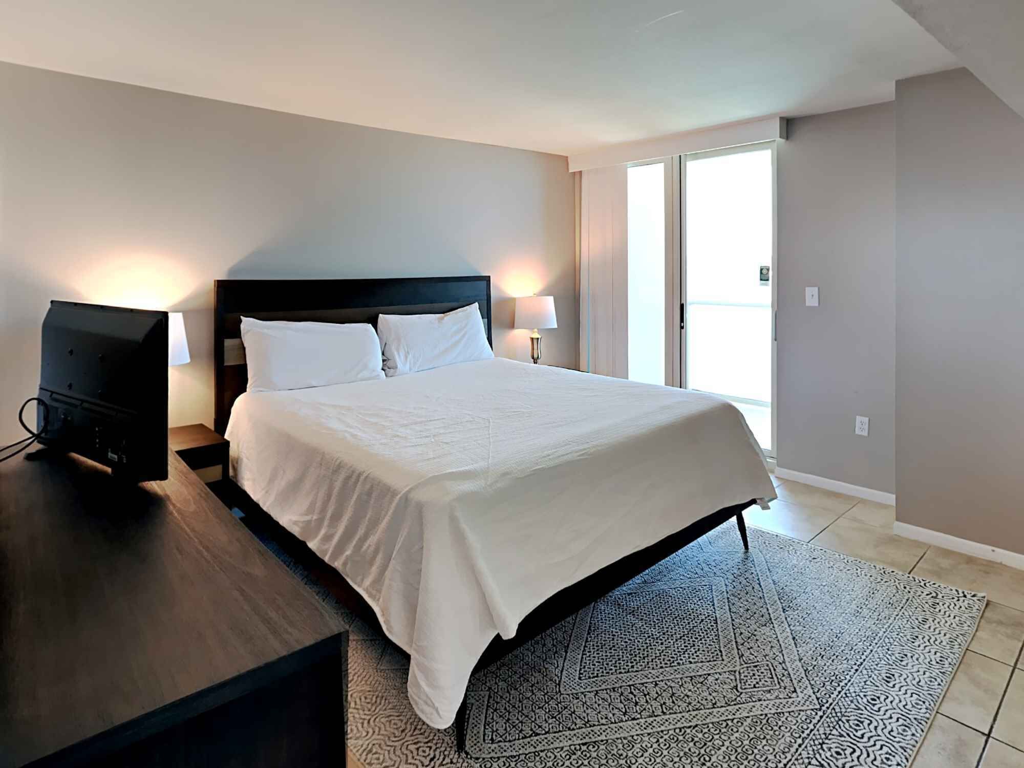 A modern hotel room with a large bed, a flat-screen TV, and a large window providing a view of the outdoors.