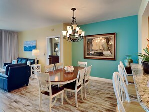 Un coin repas avec une table en bois et des chaises blanches, un lustre, un tableau représentant un voilier et un mur de couleur turquoise.