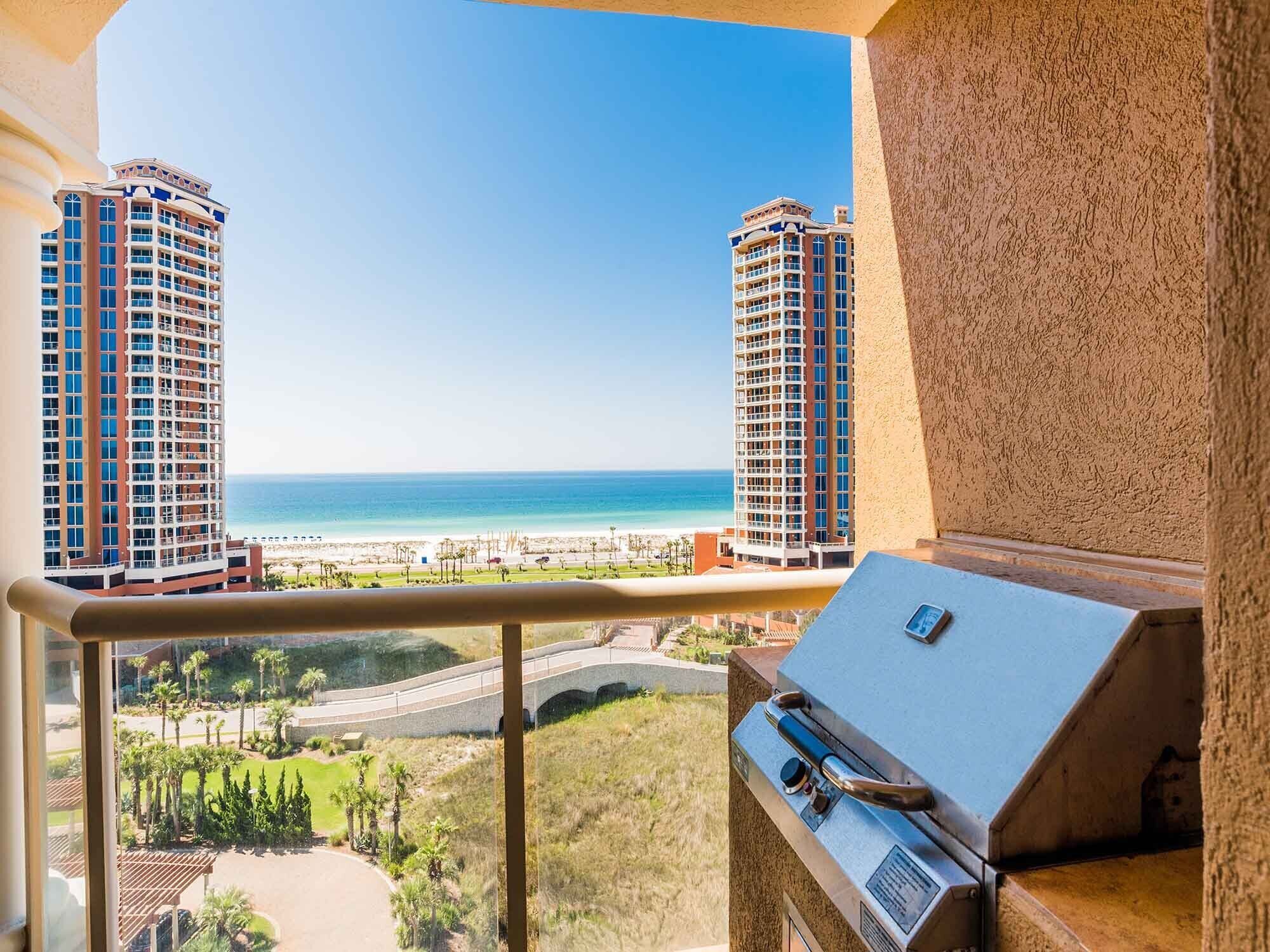 A balcony with a grill, overlooking a beach with two tall buildings and a clear sky.