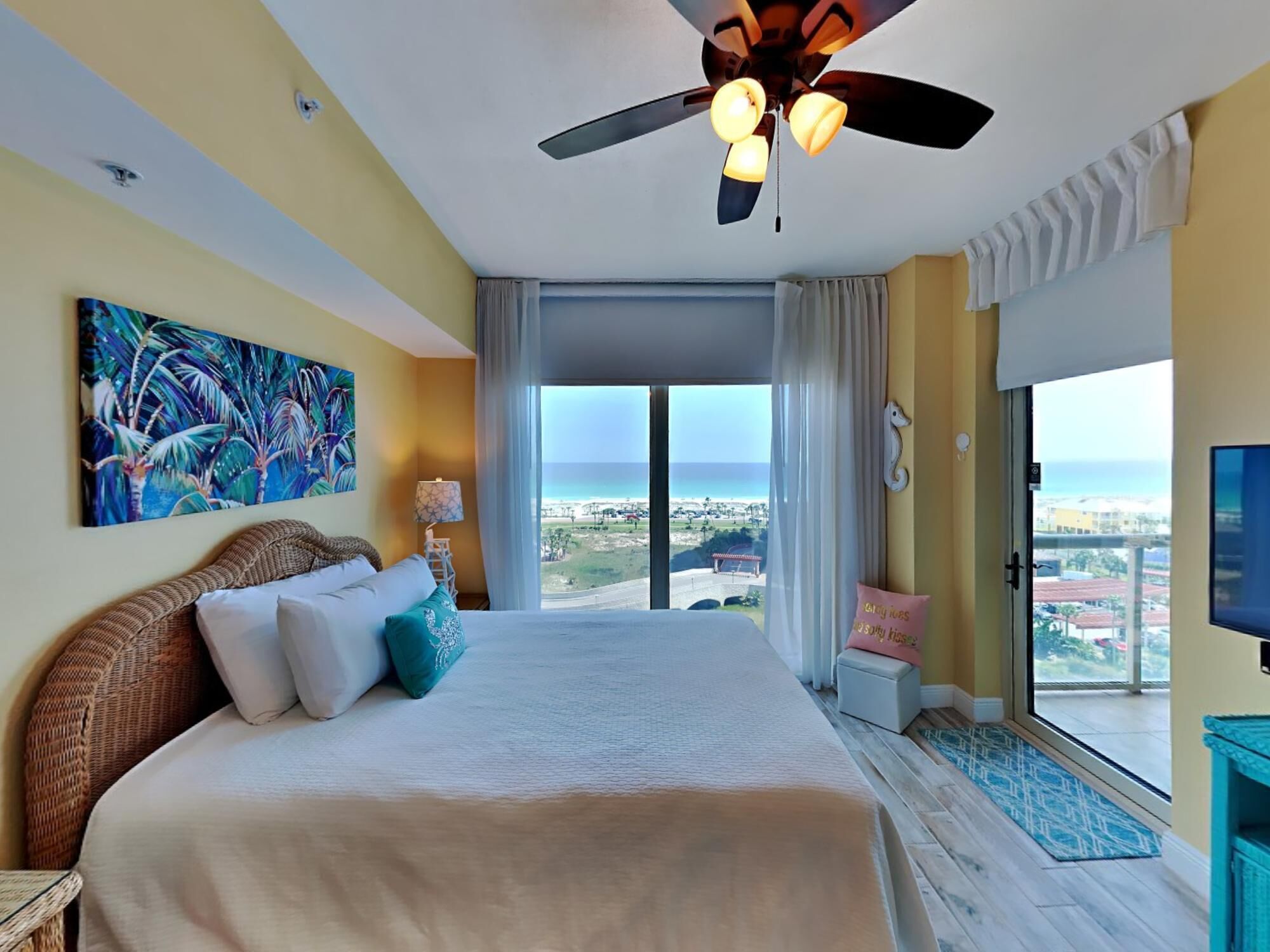 Een ruime slaapkamer met een groot bed, een plafondventilator, een schilderij, uitzicht op de oceaan en een balkon met een televisie.