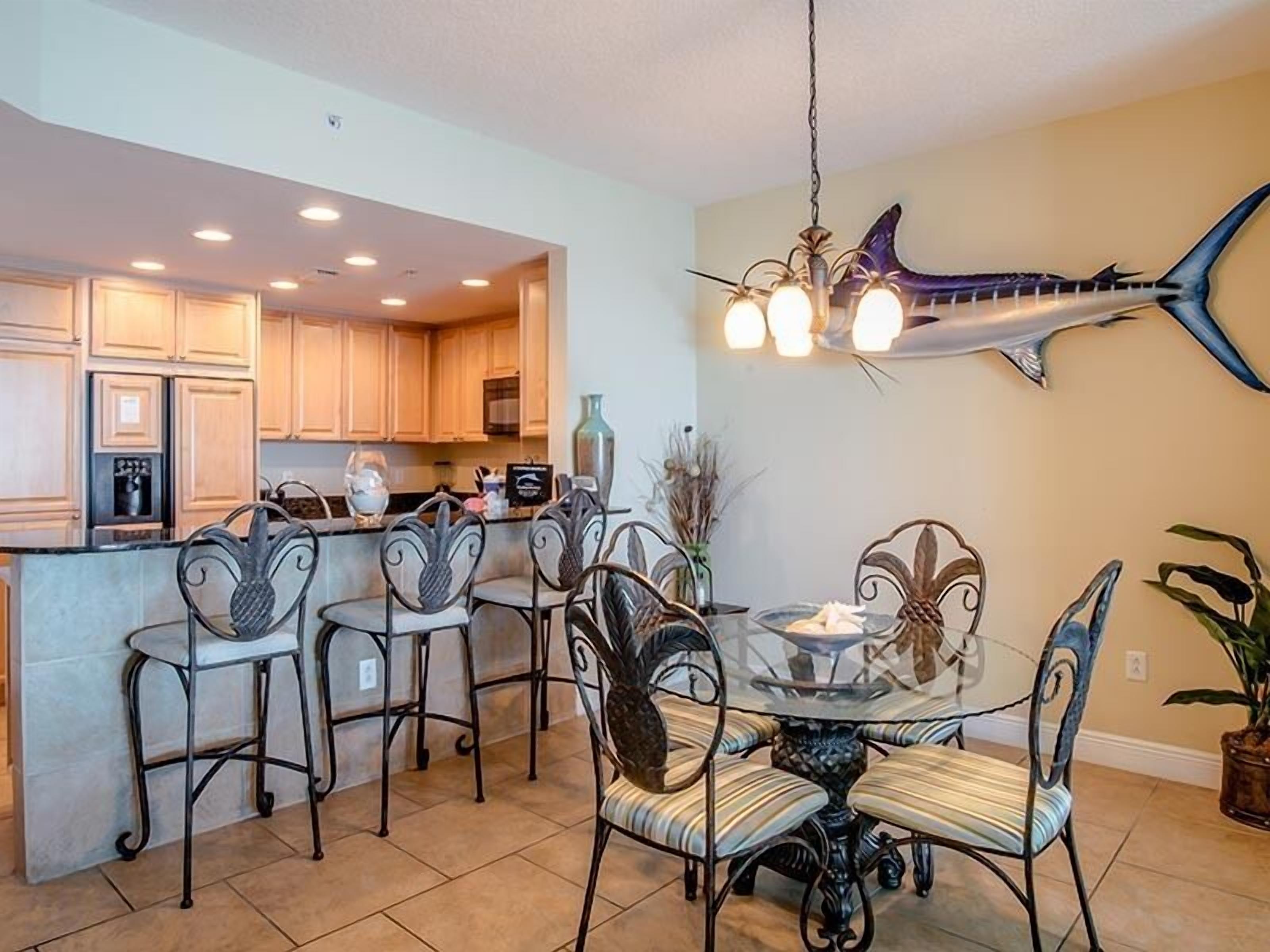 A modern kitchen with wooden cabinets, a dining area with a glass table and metal chairs, and a decorative shark mounted on the wall.