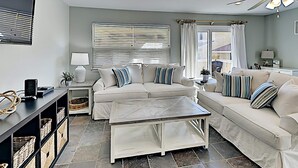 A modern living room with a beige sofa set, a coffee table, and a TV on a stand. There are wicker baskets on shelves and a ceiling fan. - Regency Cabanas by Southern Vacation Rentals (Pensacola Beach)