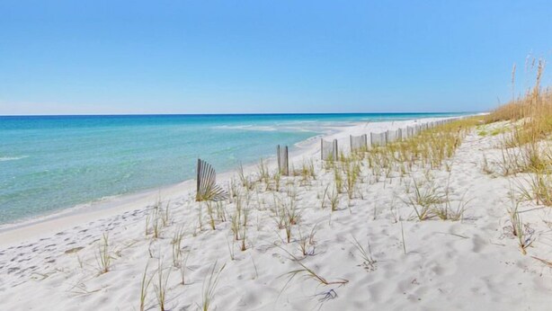 A beach with clear blue water, sandy shore, and sparse vegetation. - Regency Cabanas by Southern Vacation Rentals (Pensacola Beach)