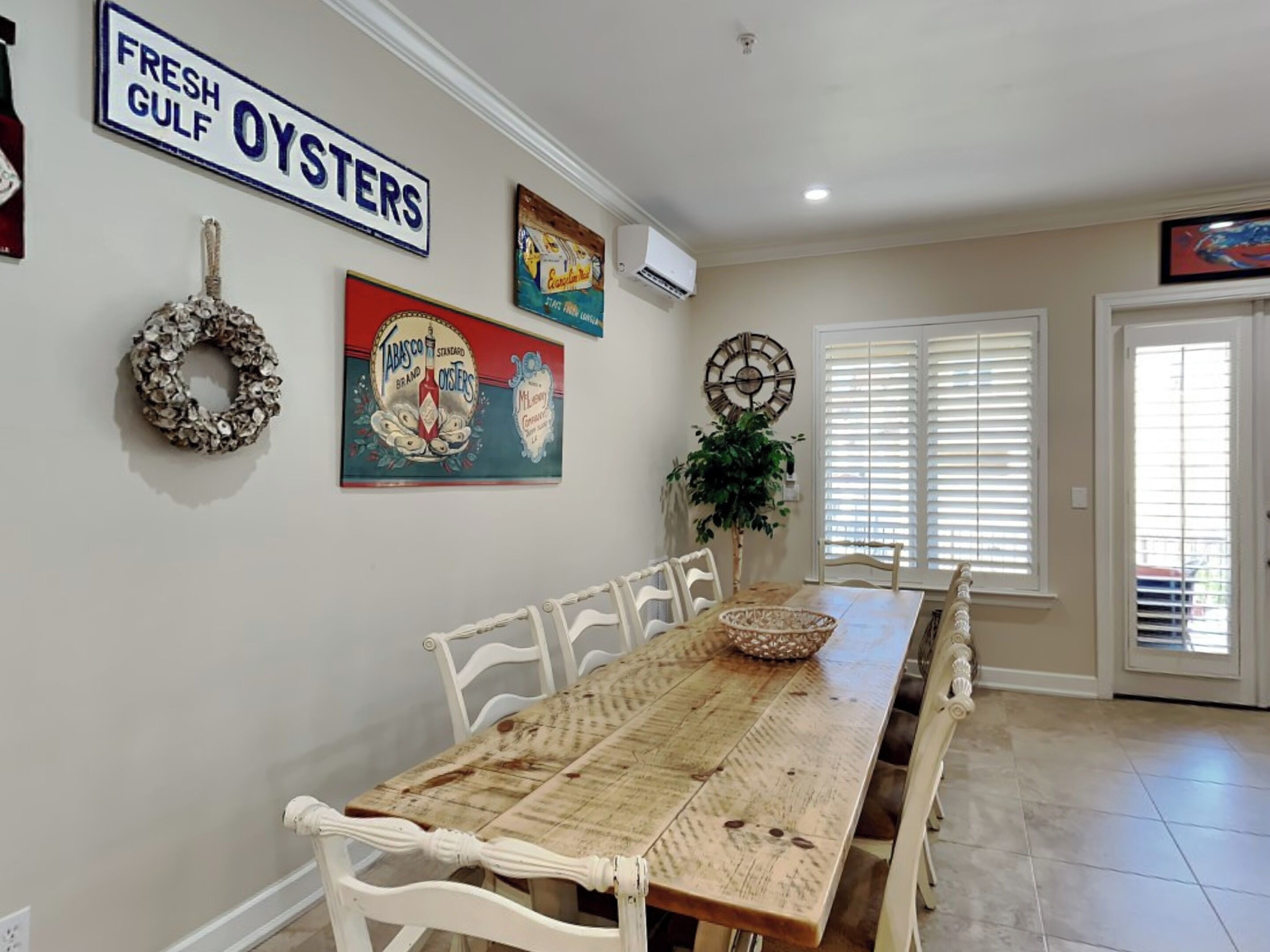 A dining area with a long wooden table, white chairs, a decorative wreath, and wall art including a sign for 'Fresh Gulf Oysters'.