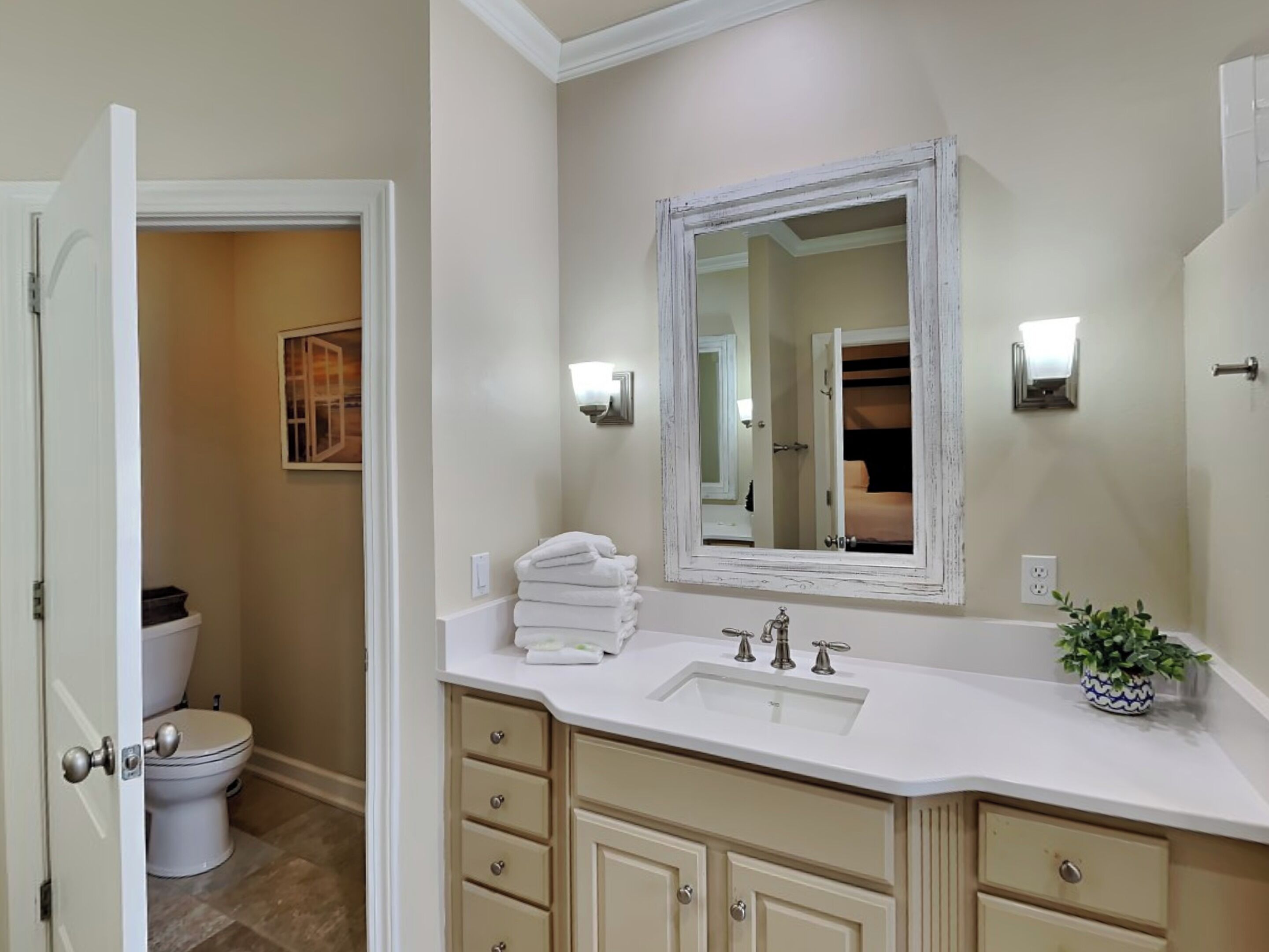 A bathroom with a white sink, vanity, and a mirror. There is a toilet visible in the adjacent room.
