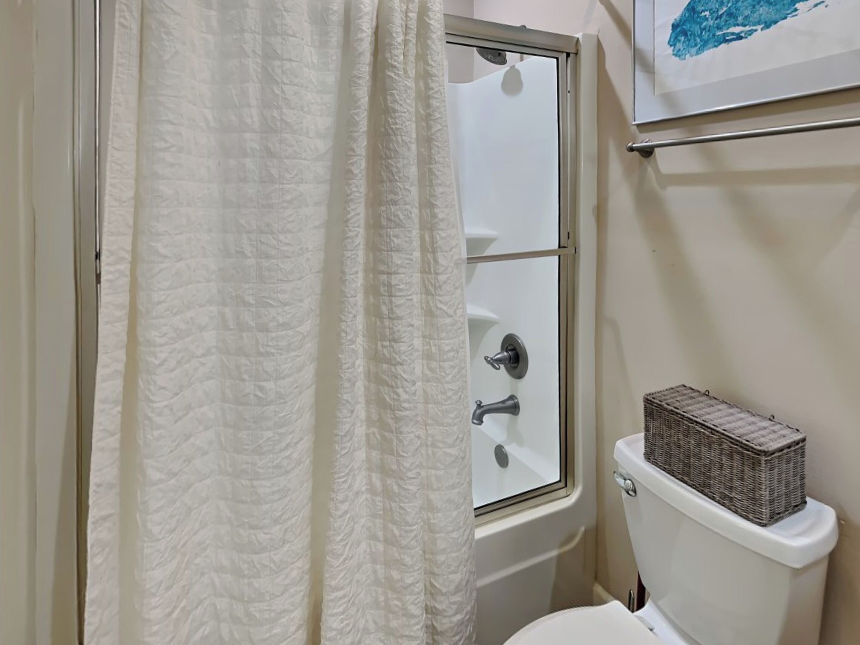 Une salle de bain avec des toilettes blanches, une fenêtre avec un rideau blanc et un petit panier posé sur le réservoir des toilettes.