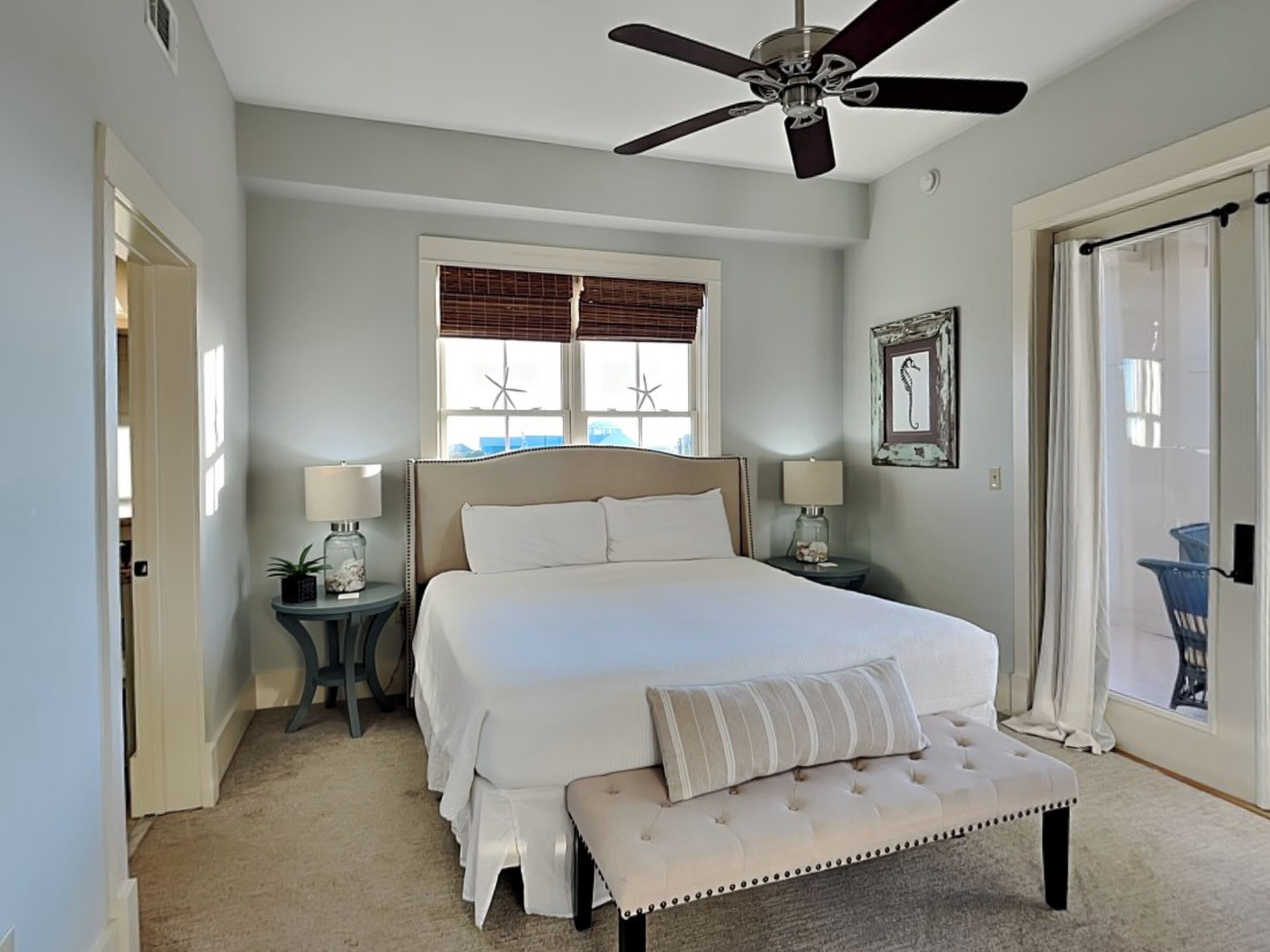 A bedroom with a large bed, two bedside tables with lamps, a ceiling fan, a window with blinds, and a door leading to another room.