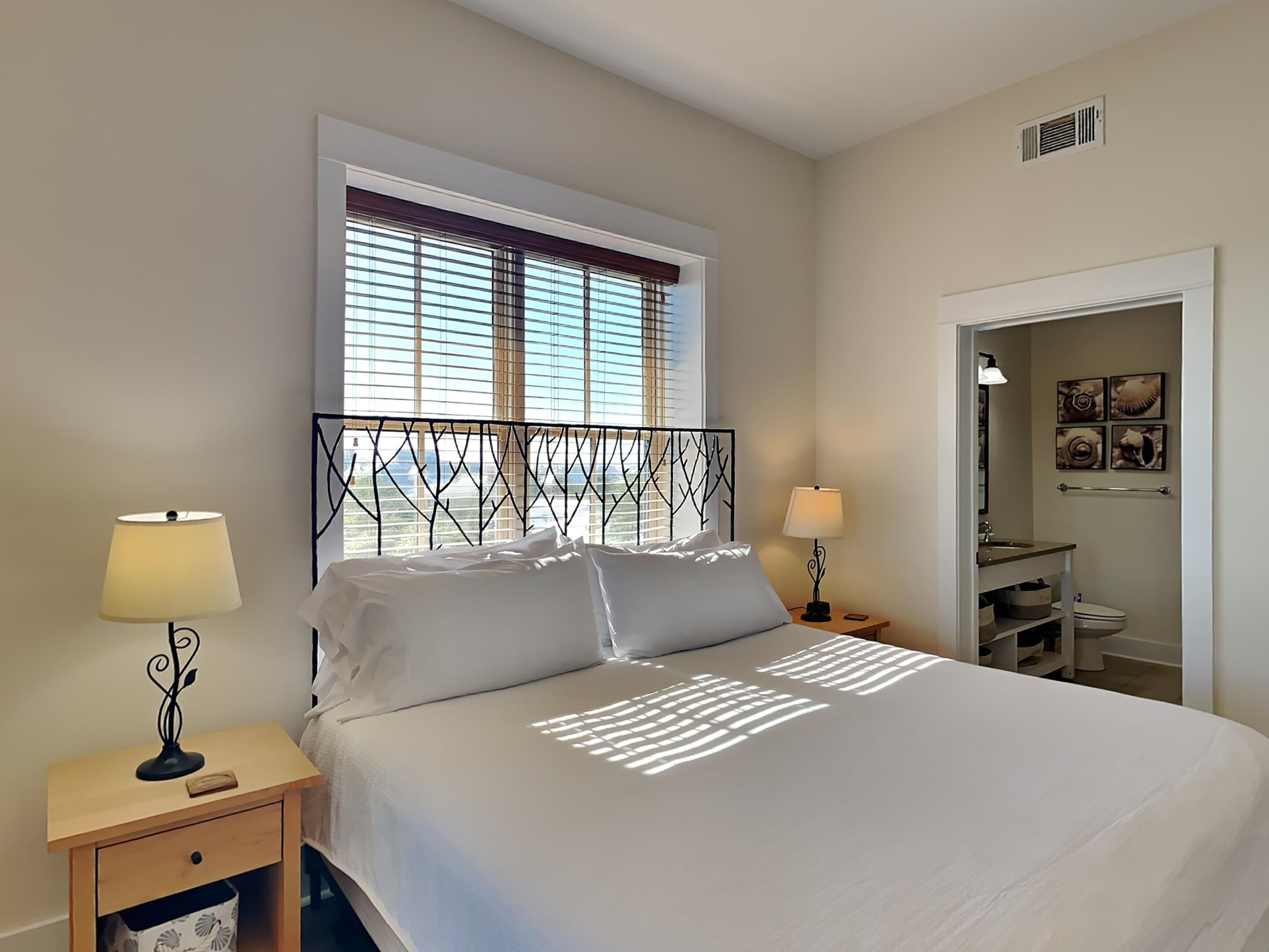 A neatly made bed with a white bedspread, two bedside lamps, a window with blinds, and a door leading to a bathroom.