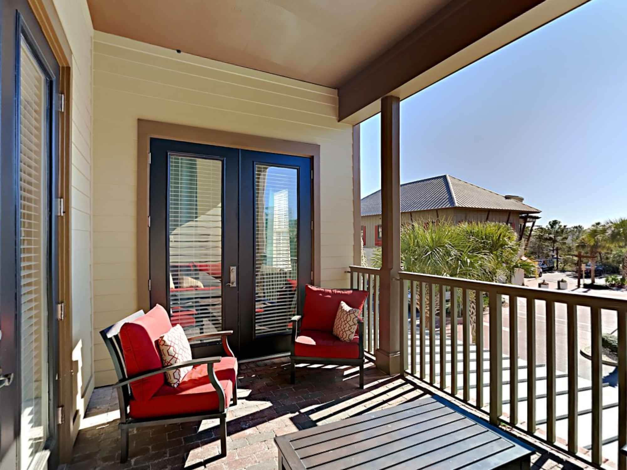 Un balcon avec deux chaises rouges et une petite table, une porte-fenêtre coulissante, et une vue sur un immeuble et des palmiers.
