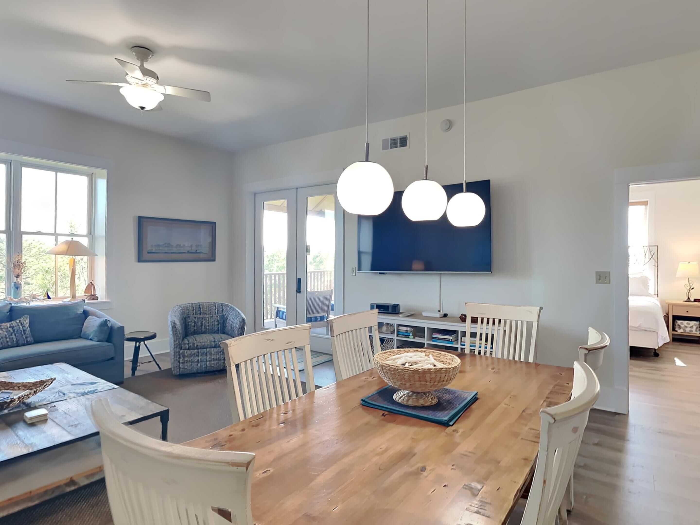 A modern living room with a wooden dining table, white chairs, a TV mounted on the wall, and a ceiling fan.