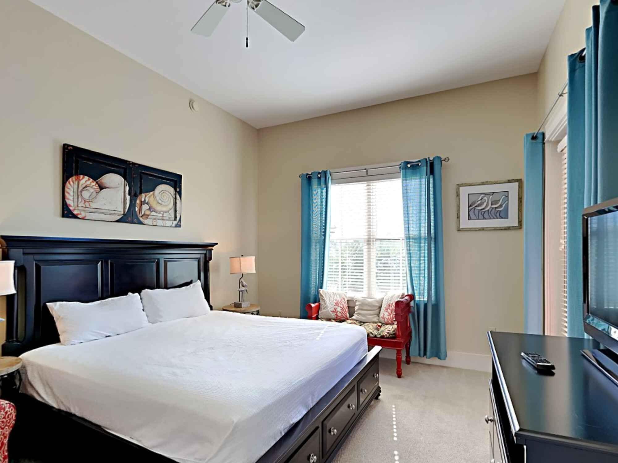 Een slaapkamer met een groot bed, nachtkastje, stoel, kapstok, plafondventilator en een raam met gordijnen.