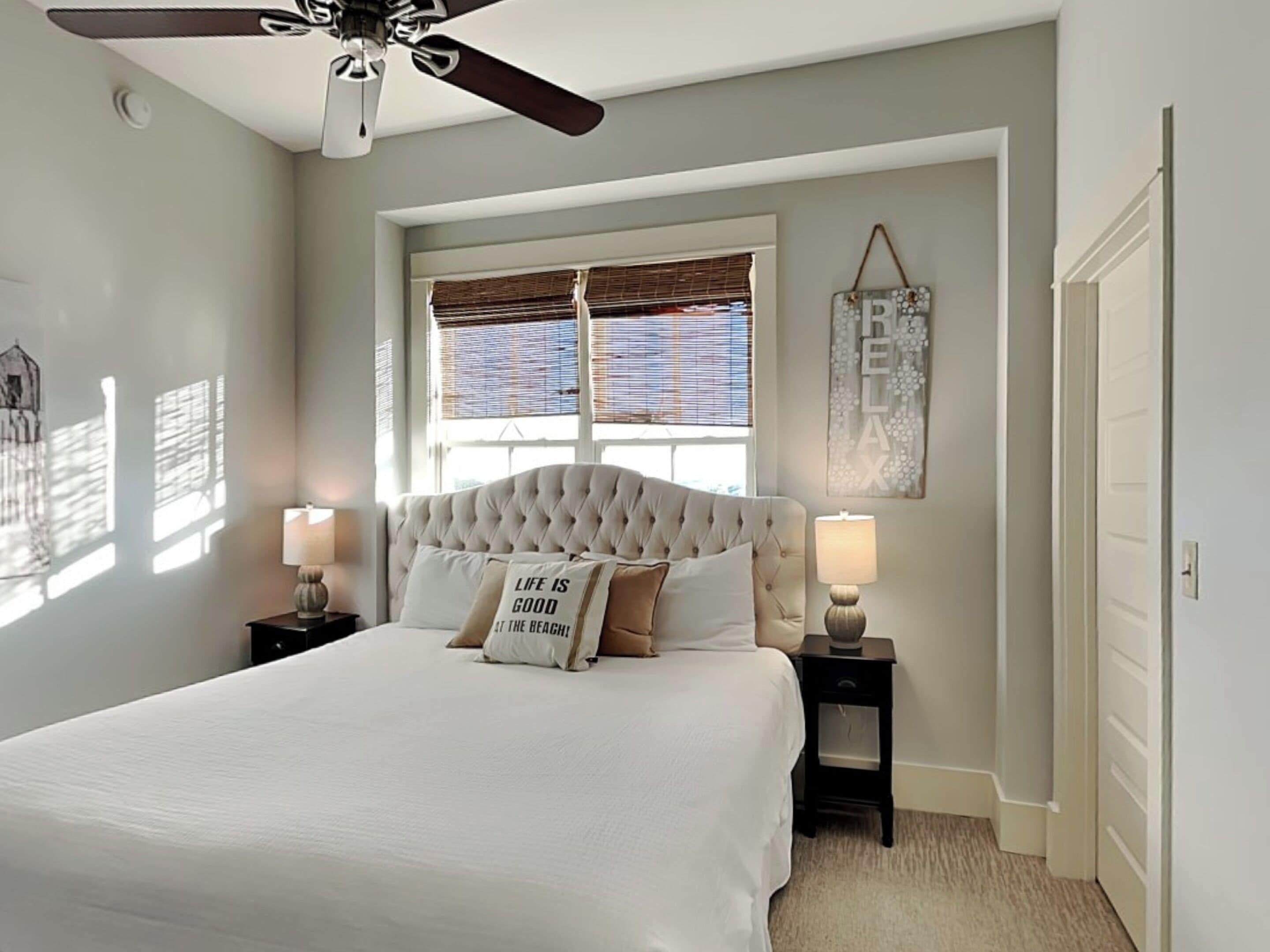 A bedroom with a large bed, two bedside tables with lamps, a ceiling fan, a window with blinds, and a door.