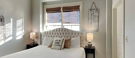 A bedroom with a large bed, two bedside tables with lamps, a ceiling fan, a window with blinds, and a door.