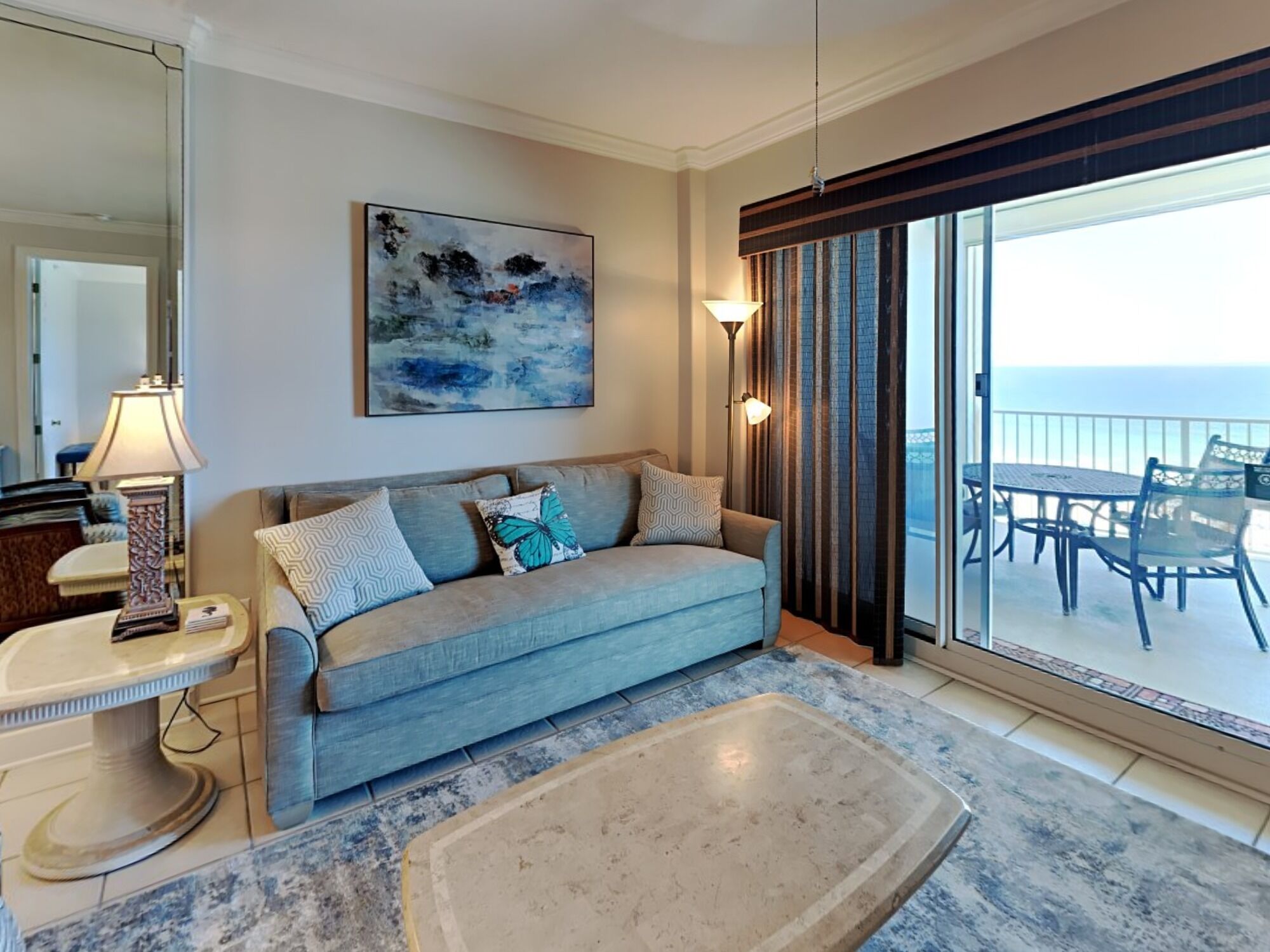 A living room with a sofa, a coffee table, a lamp, a painting, and a view of the ocean through a sliding door.