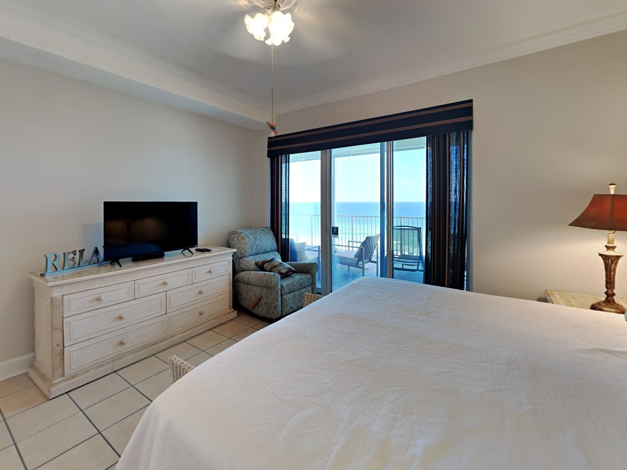 Een slaapkamer met een groot bed, een televisie op een dressoir, een stoel en uitzicht op de oceaan door een schuifdeur.