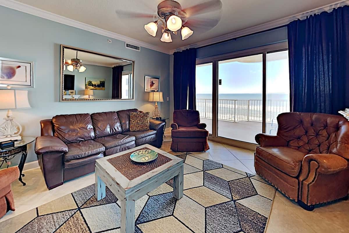 A living room with a brown leather sofa, a matching armchair, a coffee table, a ceiling fan, a large mirror, and a view of the ocean through a sliding glass door.