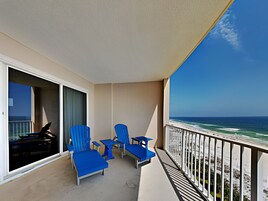 A balcony with blue chairs and a table, overlooking a beach with waves.