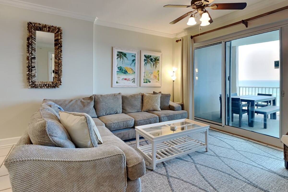 A living room with a sofa, a coffee table, and a ceiling fan. There are framed pictures on the wall and a view of the outside through a sliding glass door.