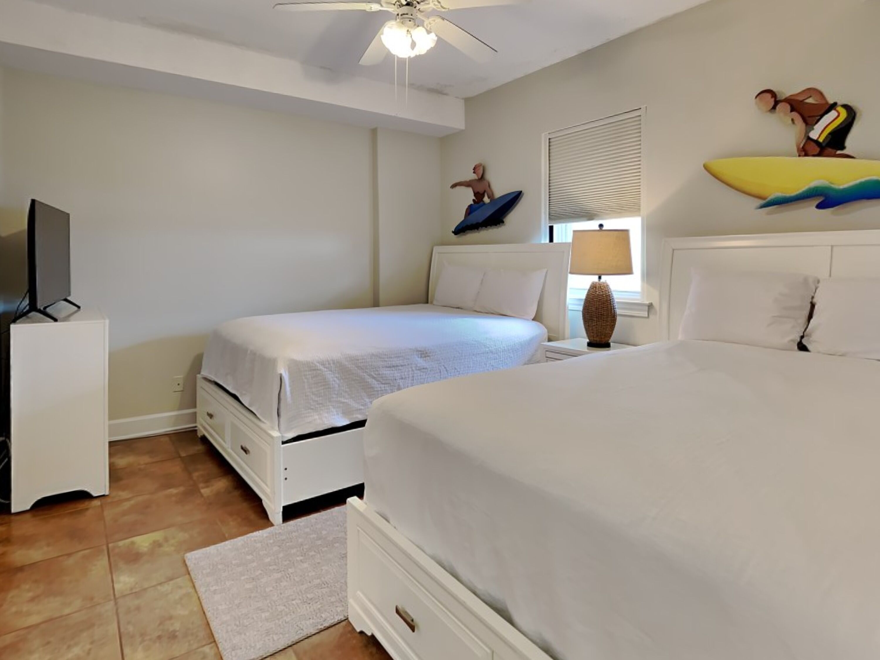 Een hotelkamer met twee bedden, een plafondventilator, een televisie op een standaard en een raam met jaloezieën.