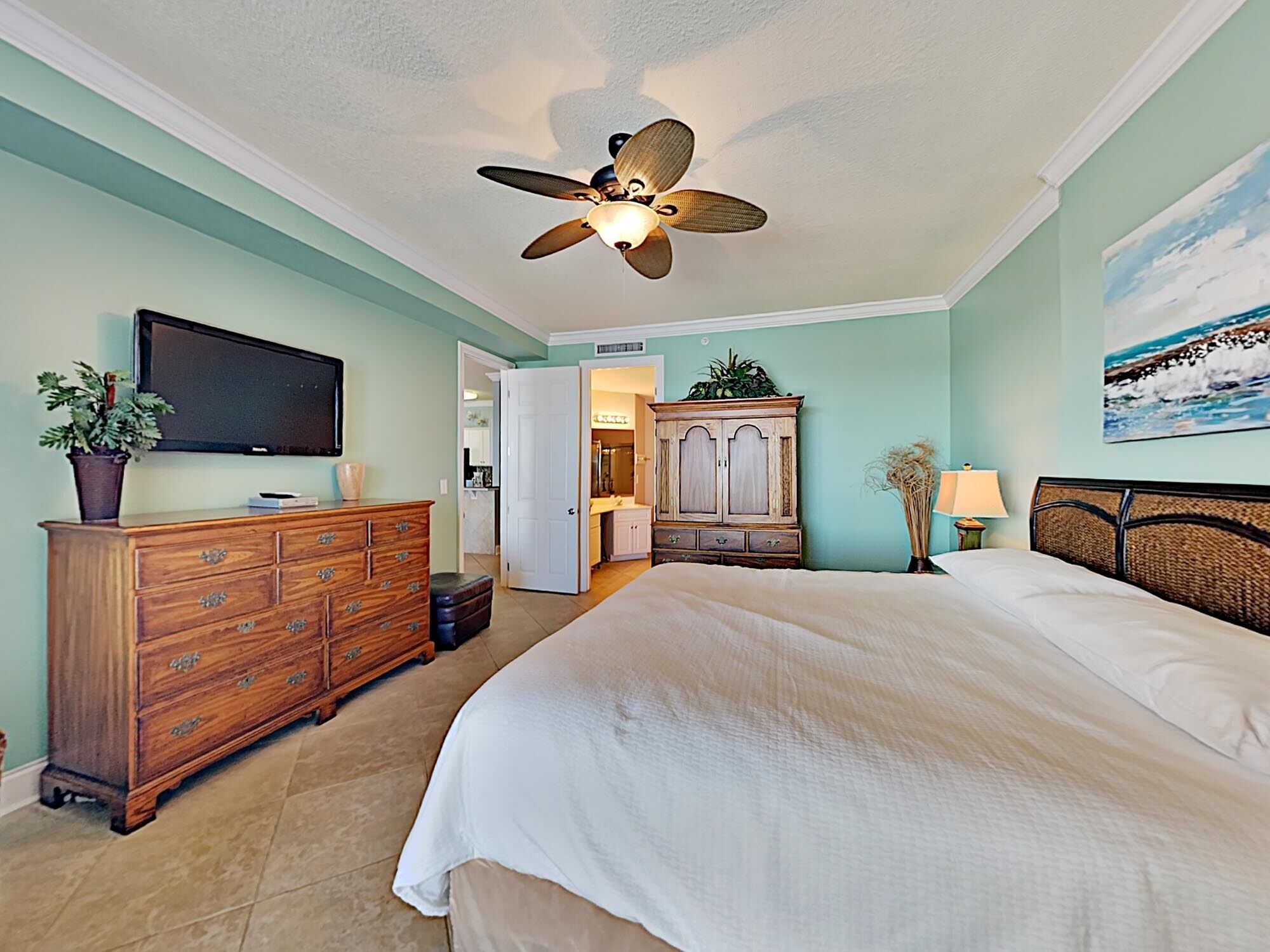 A bedroom with a bed, dresser, television, ceiling fan, and a painting on the wall.