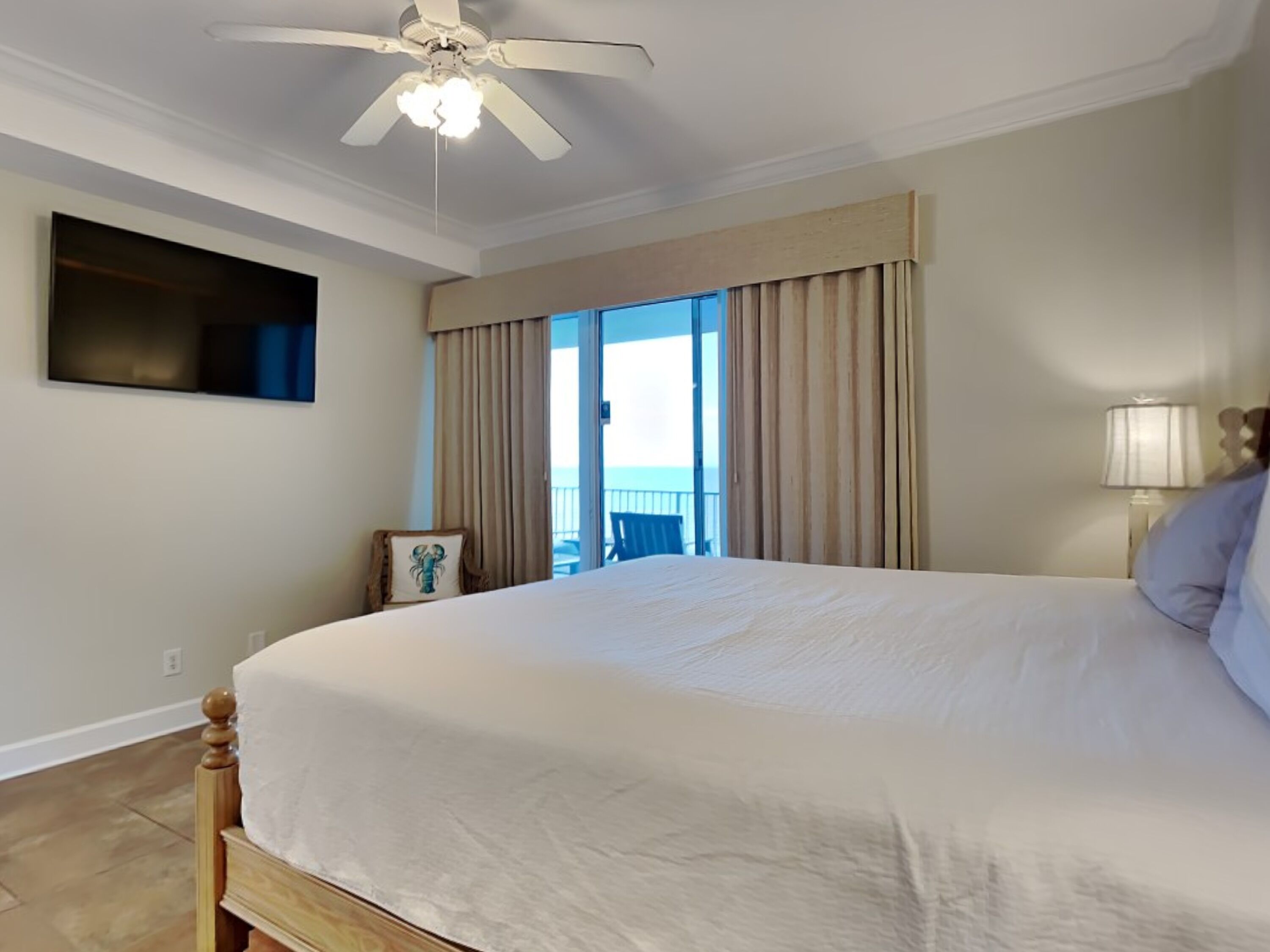 A large bed with a wooden headboard, a ceiling fan, a flat-screen TV mounted on the wall, and a view of the ocean through a sliding glass door.