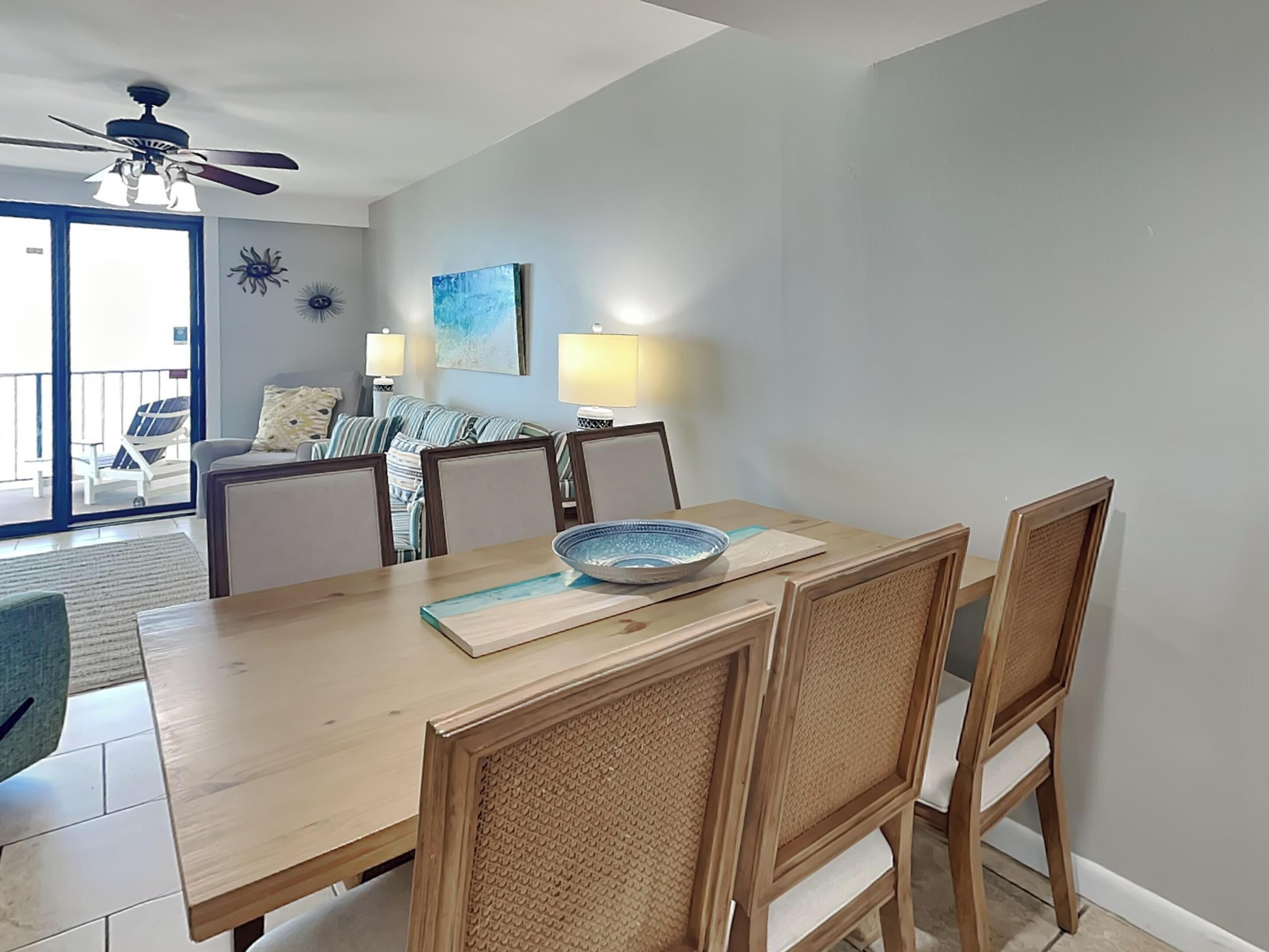 Un coin repas avec une table en bois, des chaises et un saladier. Un ventilateur de plafond est installé au-dessus, et on aperçoit un balcon avec une chaise à l'extérieur.