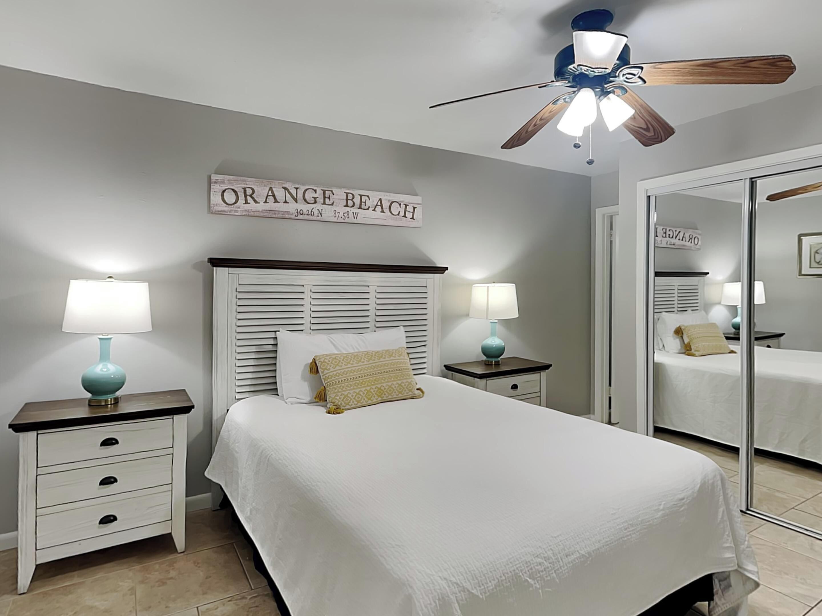 A bedroom with a bed, two bedside tables with lamps, a ceiling fan, and a sign on the wall.