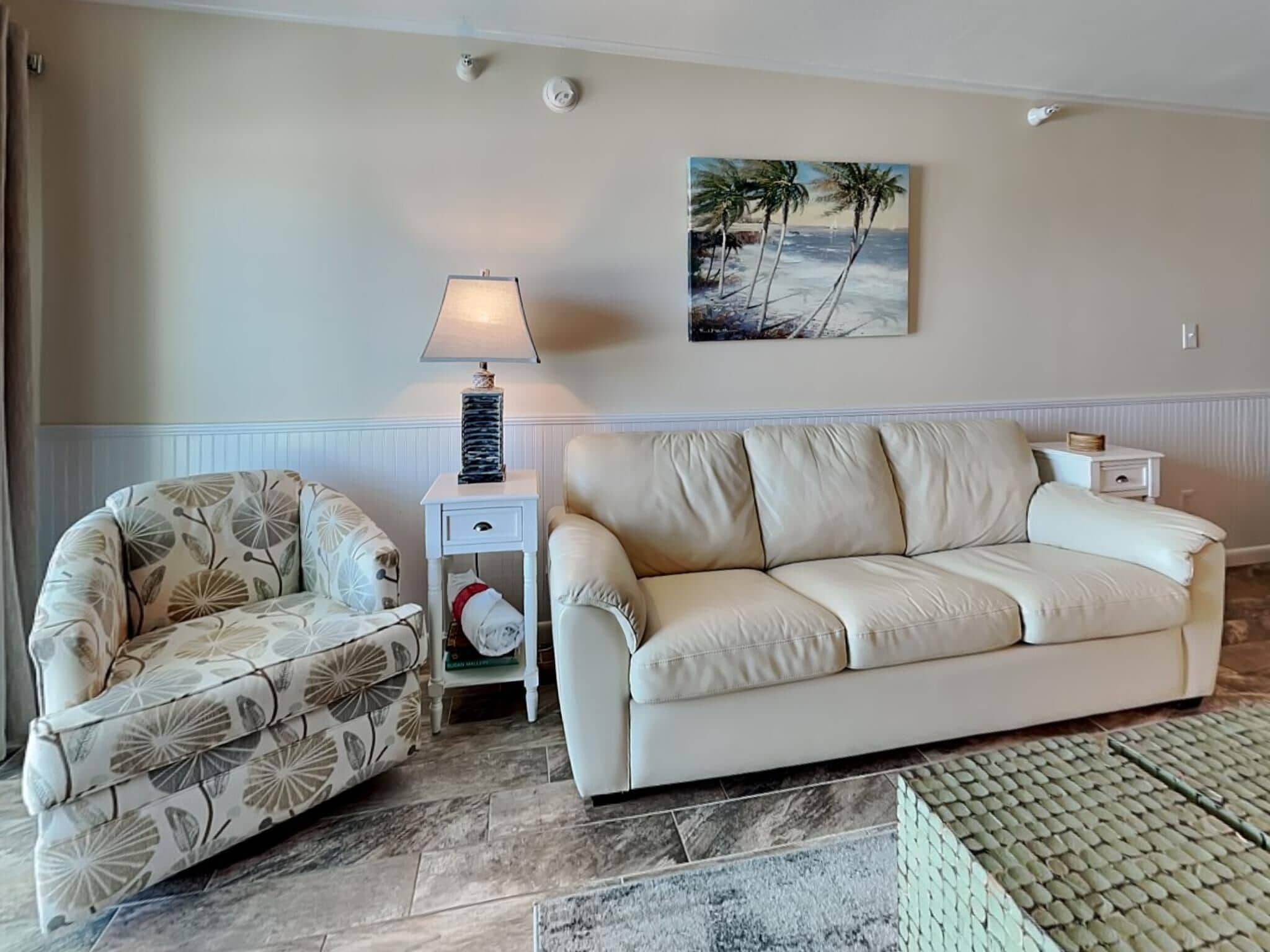 A living room with a beige sofa, a patterned armchair, a small table with a lamp, and a beach-themed painting on the wall.