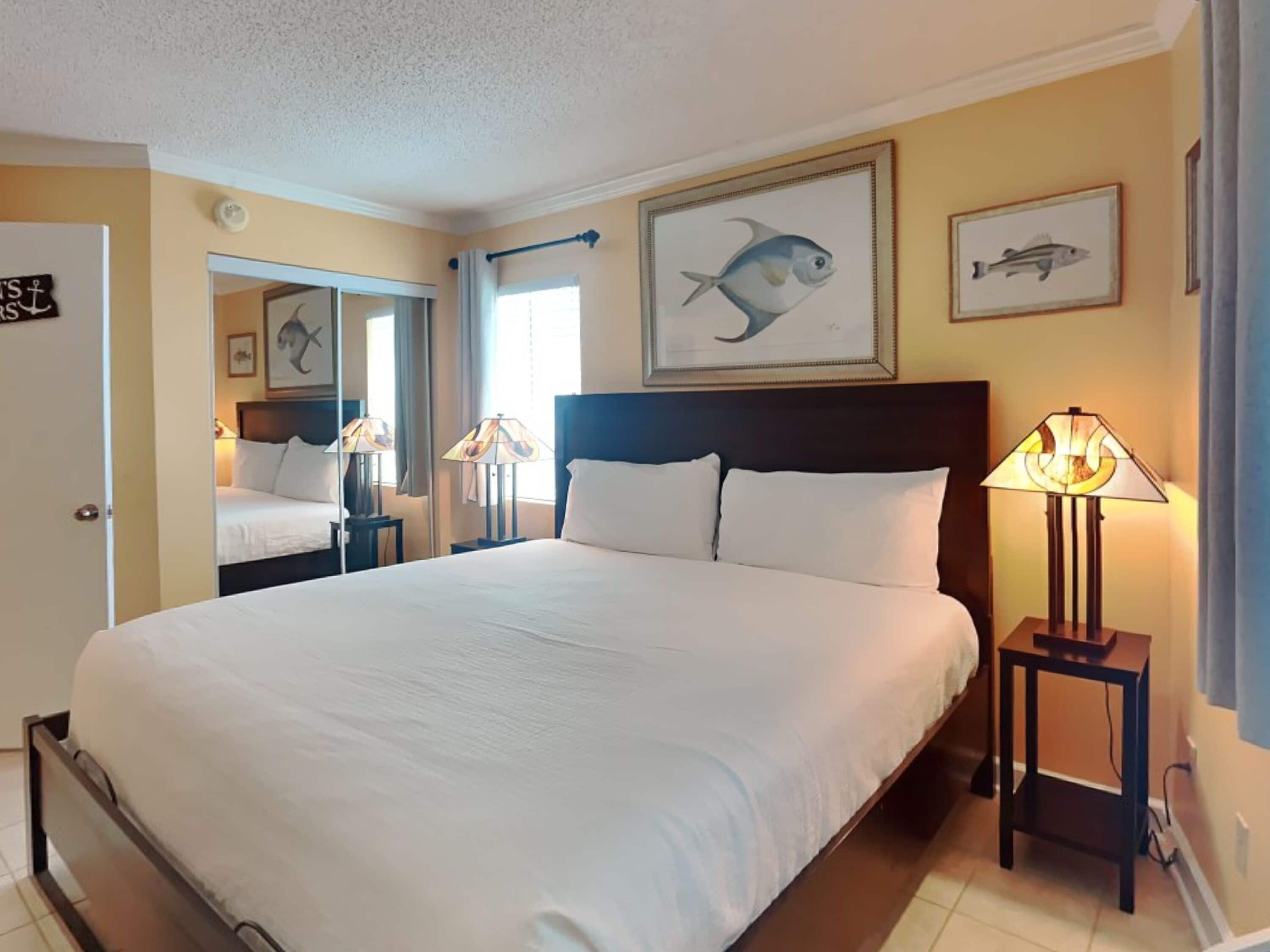 Habitació d'hotel amb un llit gran, dues taules de nit amb làmpades, un mirall i obra d'art de peixos emmarcats a la paret.